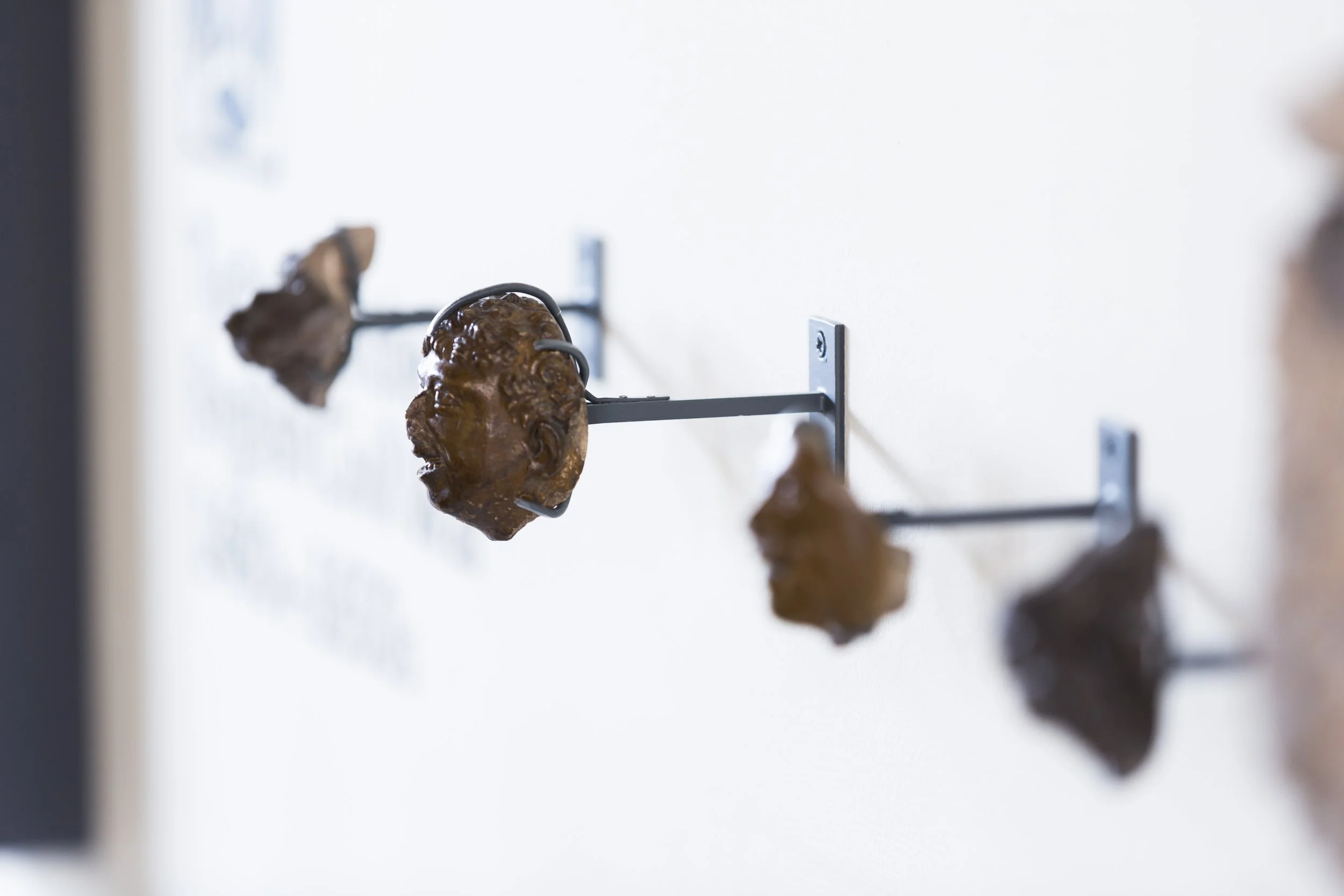 A close up of a row of small brown artefacts mounted on a white wall. Only the second object from the left is in focus: a carved head with slightly monstrous human features, an open mouth and curly hair.