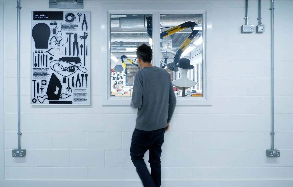 A man looks through an internal window at a workshop space in the background. To his left is an artwork labelled "Milinery & Jewellery" featuring silhouettes of industry tools.