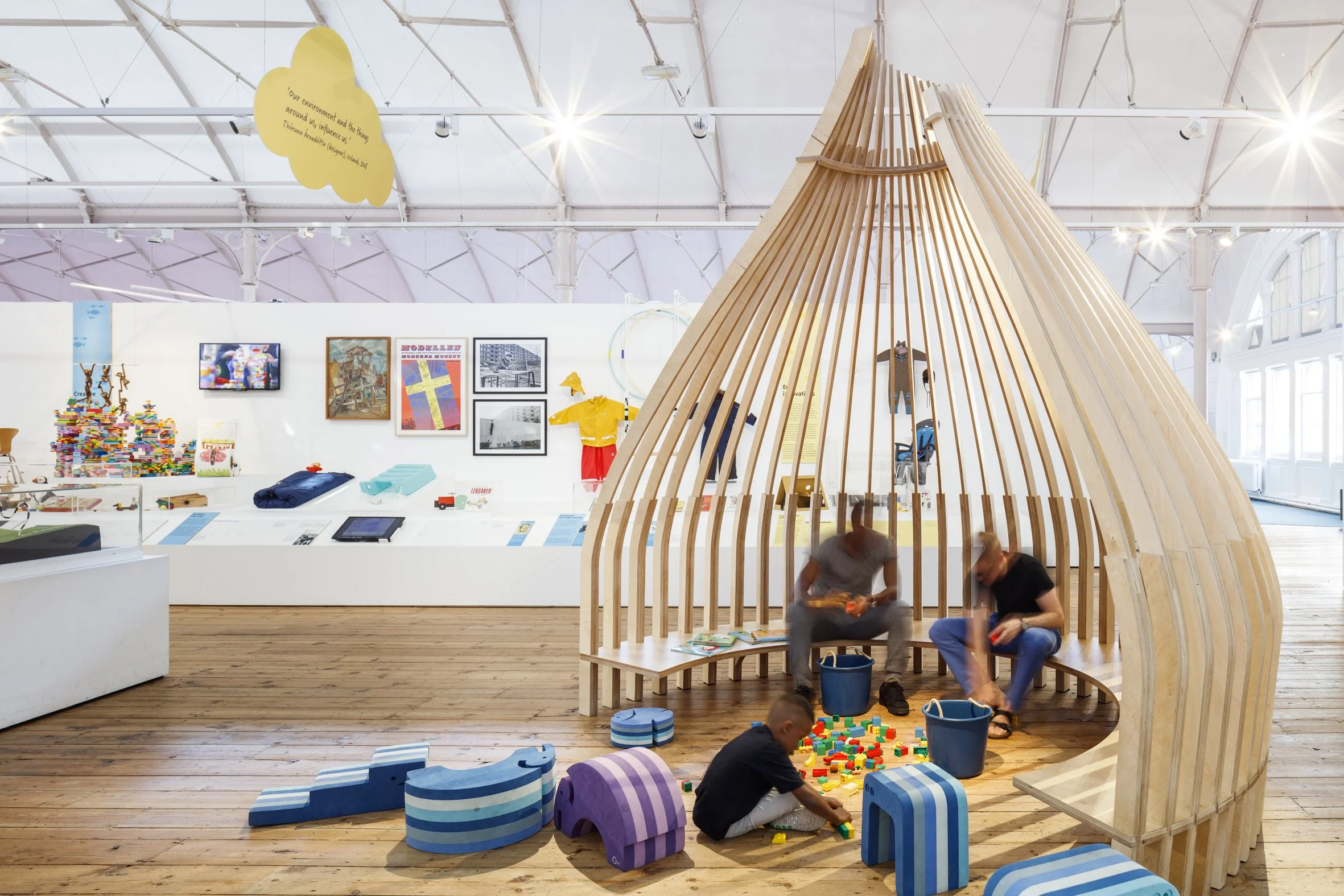 A minimal timber frame in the shape of a rounded hut, open on one side with a low bench around its inner edge. Two adults on the bench and a child sitting on the floor play with colourful toy building bricks in blue buckets in front of them.