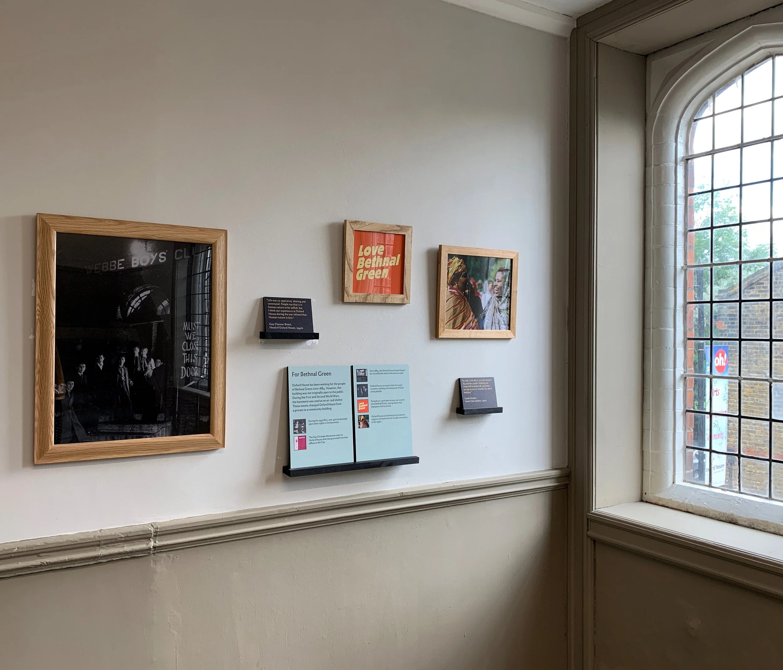 Three framed images hang in the corner of a room next to a paned window: an old photo of a boys club, a red sign saying “Love Bethnal Green”, and a colour photo of two smiling Somali women. Text labels on the wall provide more information.