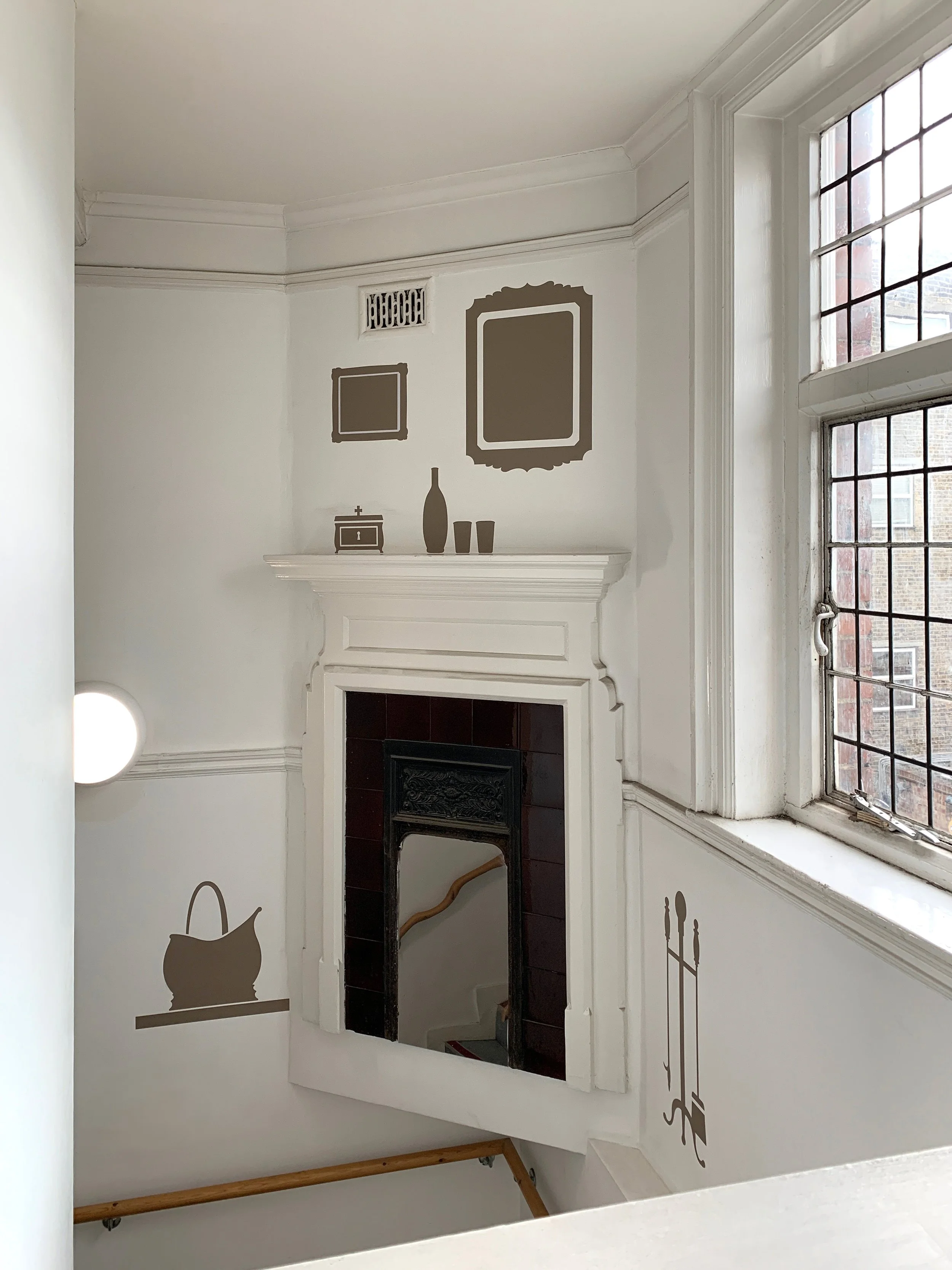 A disused fireplace halfway up a wall at the corner of a staircase. It is surrounded by 2D wall graphics which show items which would have surrounded it – a mirror on the wall, a vase on the mantlepiece, a coal basket.