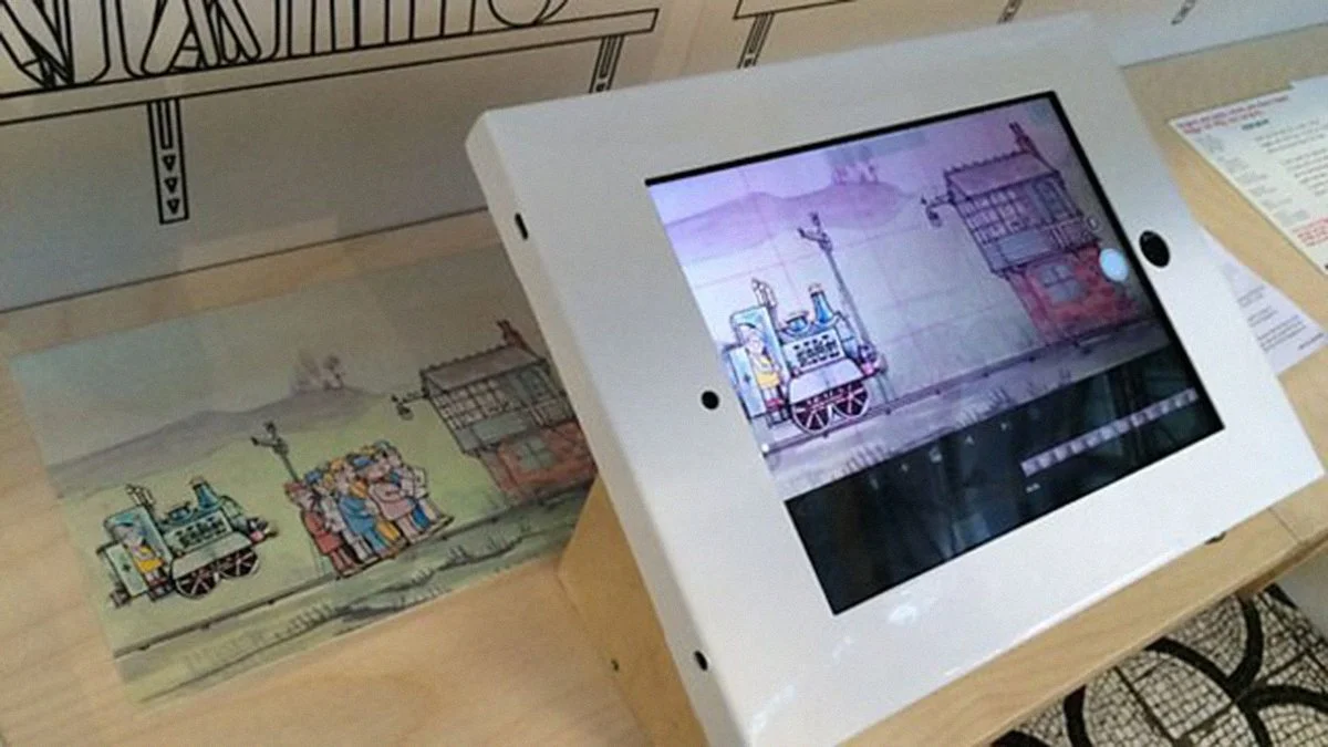 An iPad displayed at an angle on a wooden table. Beneath it is a long strip of paper with a coloured drawing of hills. The iPad screen shows the same scene with a train going by.