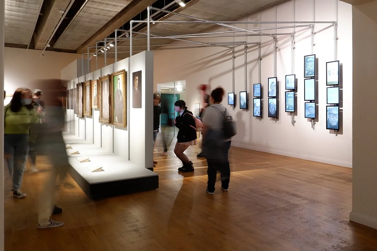 On the left of a gallery visitors look at a set of painted portraits displayed on a freestanding wall. On the back of that wall, a group of visitors take selfies. Behind them, visitor faces are displayed on digital tablets.