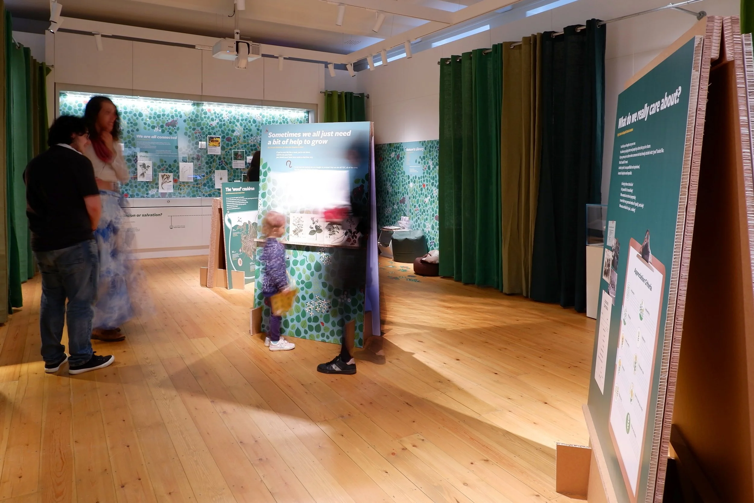 Two cardborad panels stand at the centre of the gallery. Curtains in shades of green surround the space. Graphics with leaves and natural scenes are all around. Three individuals are having conversation while a child participates in an interactivity.