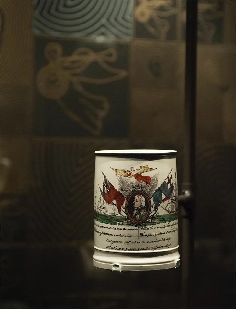 A white ceramic tankard in a glass showcase mounted on a pole with out-of-focus patterns behind it. The tankard features a painting of a man in an oval frame with an angel above him, flanked by ships and two flags.