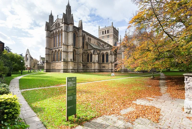 A path leading to a large church amidst green grass and trees, with older relics in the background. In the foreground, a green signboard with white text and arrows shows directions to different locations.