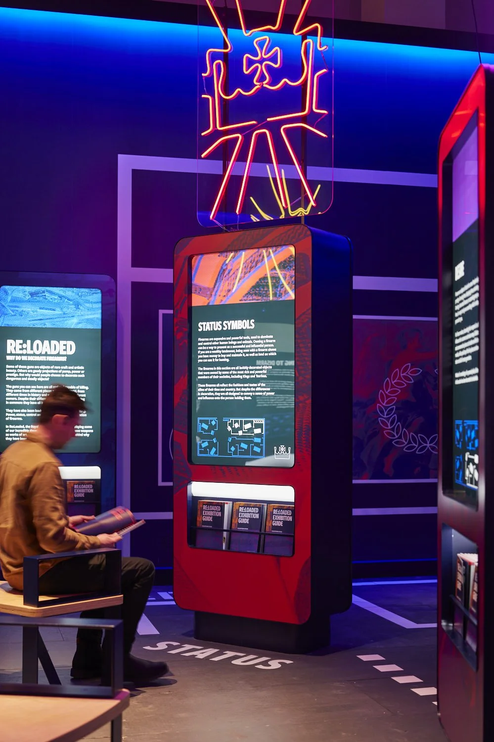 A person sits on a bench reading an exhibition guide in front of a graphic introductory panel. At the top, the text says "Status symbols," and below, a recessed aperture contains guides for visitors to read as they view the objects.
