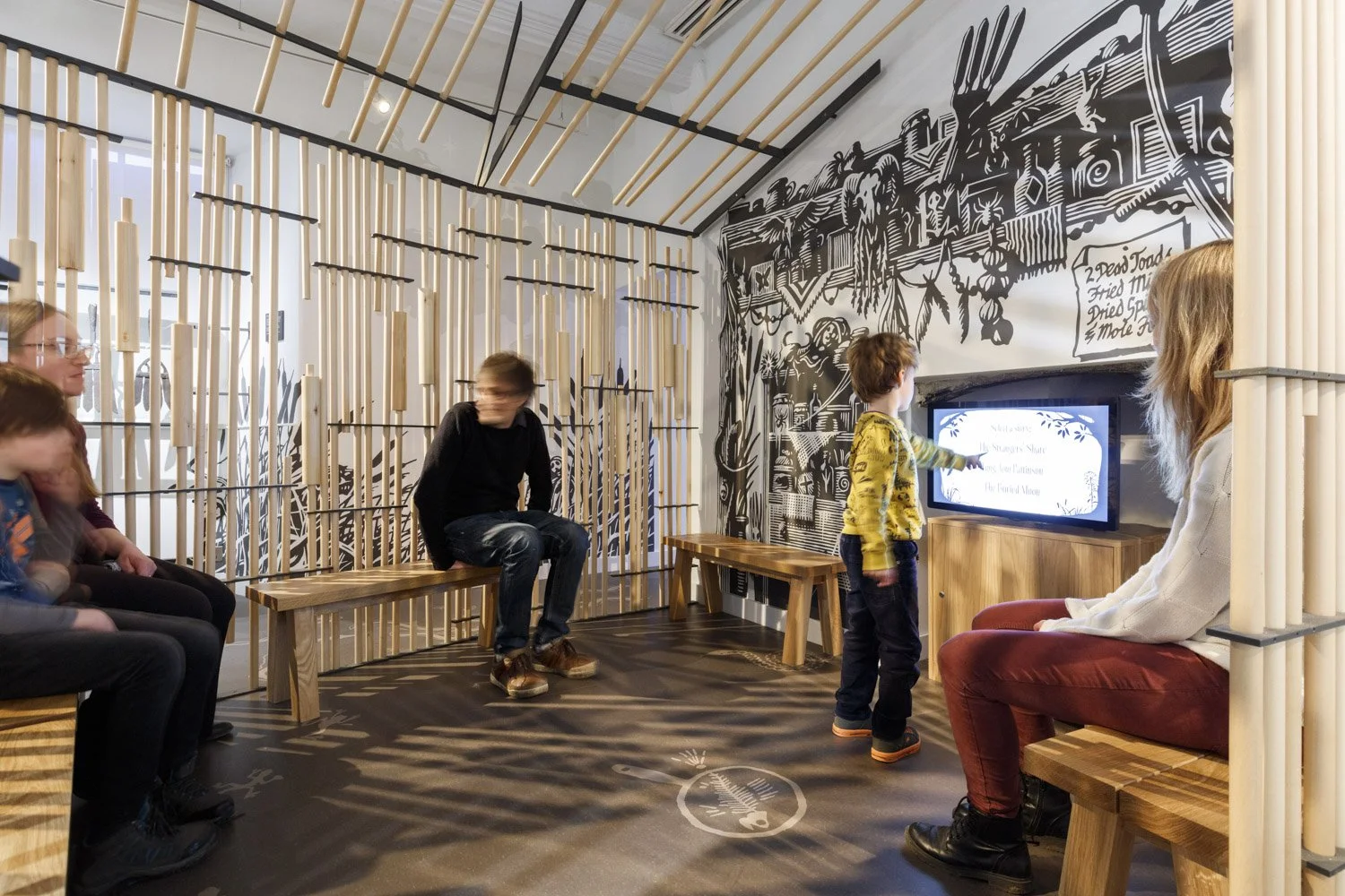 Adults and children sit on benches inside an open timber structure. There is a video display beneath a black and white artwork of traditional herbalist equipment, including bottles and a ram’s skull.