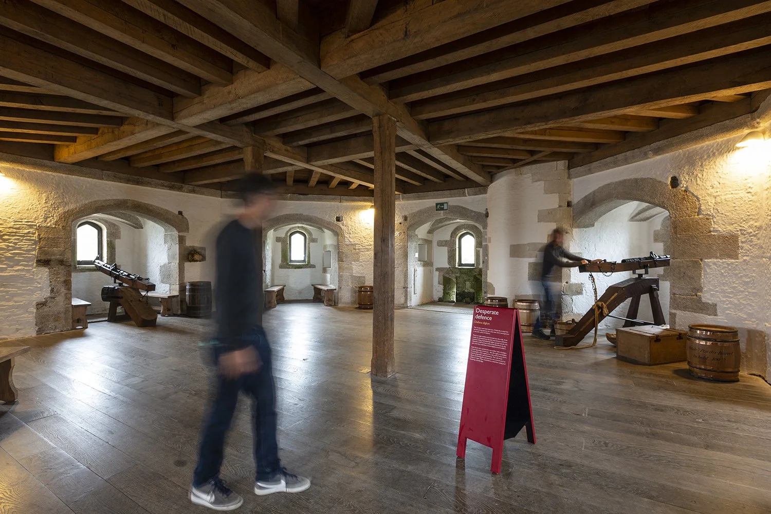A circular room with rough whitewashed stone walls, wood beamed ceiling, and window openings in recessed alcoves. A visitor plays with one of two canons positioned at the windows. In front, a man walks by a red A-frame sign titled “Desperate defence”