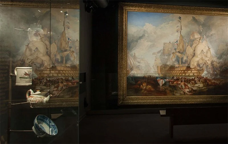 A painting of a ship in a gilded frame in a dark room. To the left, the painting is reflected in a glass showcase. Inside the showcase are four ceramic pieces suspended from a central mount, including a bowl, teapot and tankard.