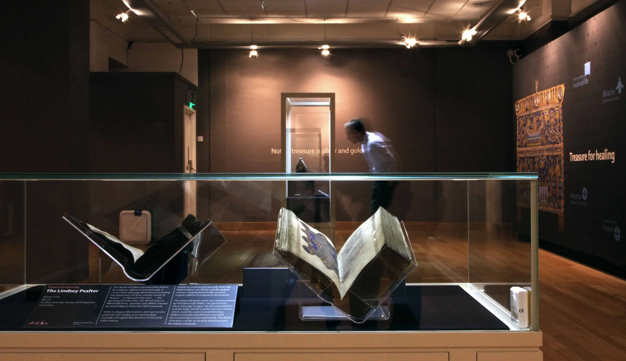Two open manuscripts in a spotlit showcase. In the background, a visitor looks at objects in another showcase.