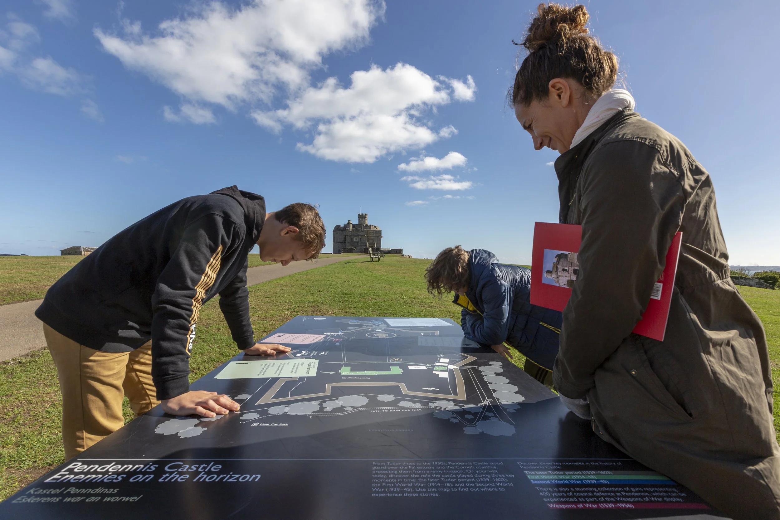 A woman and two young people stand around a large horizonal sitemap titled ""Pendennis Castle: Enemies on the Horizon"". Behind them, a path leads up to the castle under a sunny sky.