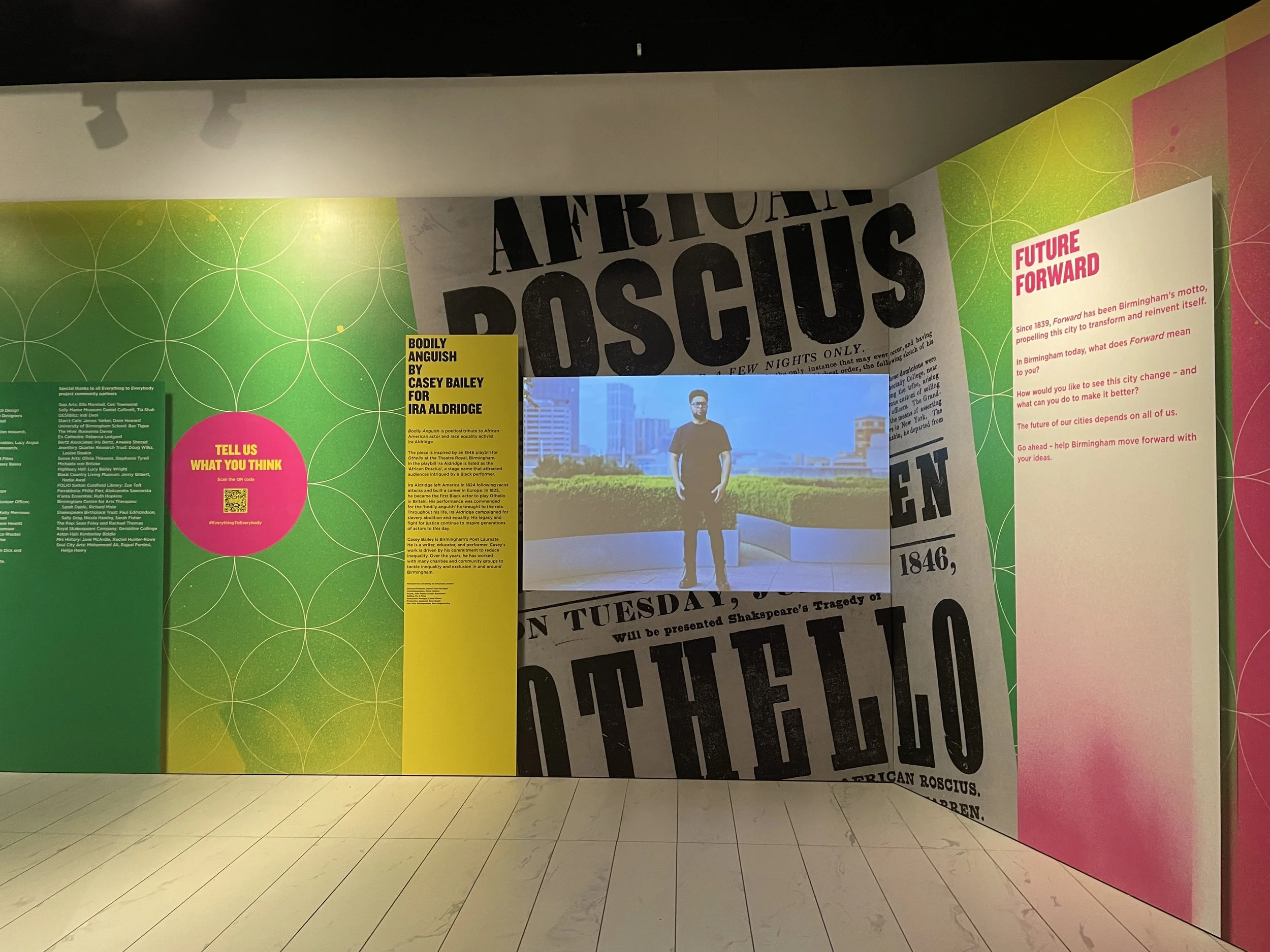 A video of a man with a cityscape behind him is projected against a colourful gallery wall with a green, yellow and pink spray-paint effect and a flyer for Shakespeare’s Othello. The video is titled “Bodily Anguish by Casey Bailey for Ira Aldridge”