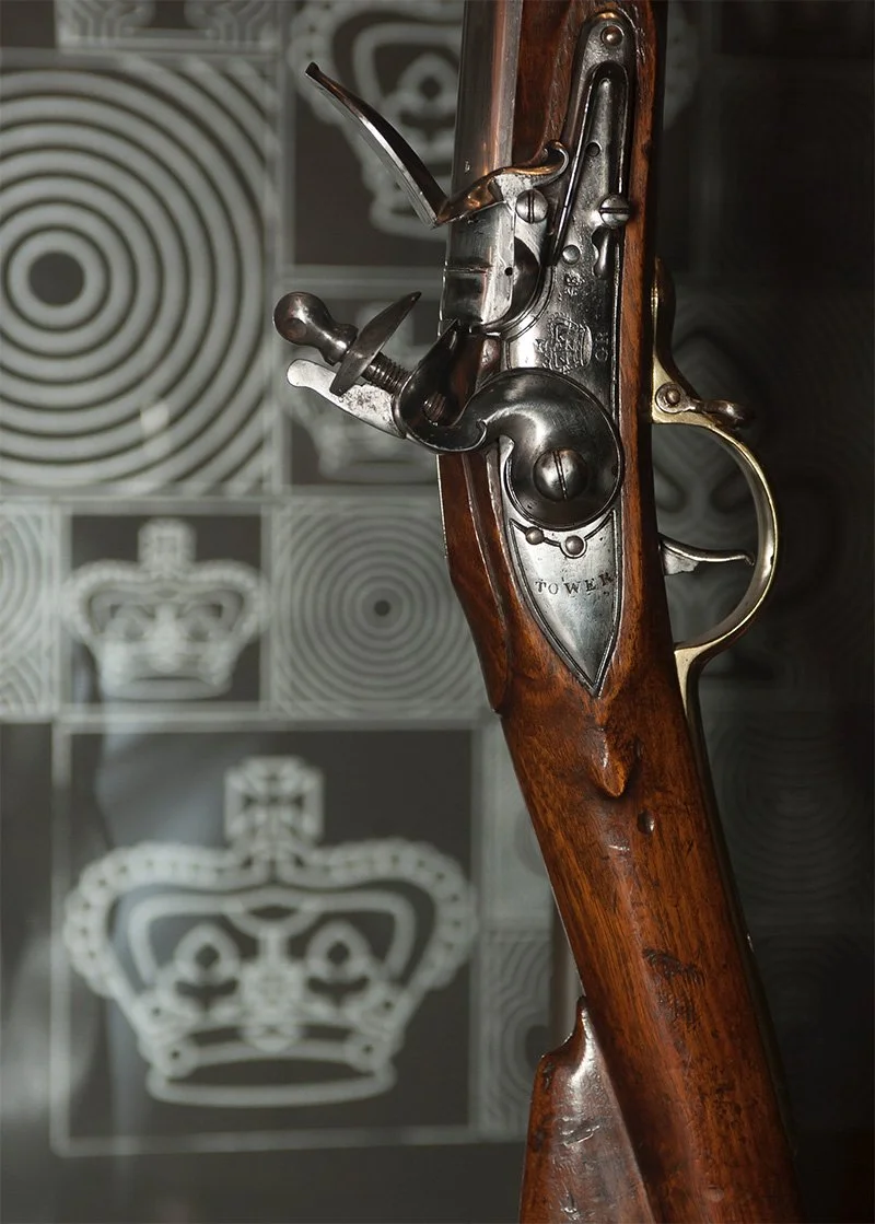 A close up of the trigger of a wooden long-barrelled gun, with a series of polished metal fittings. The word “tower” and a crown symbol are etched in the metal. Behind the gun are out-of-focus motifs of crowns and circular lines.