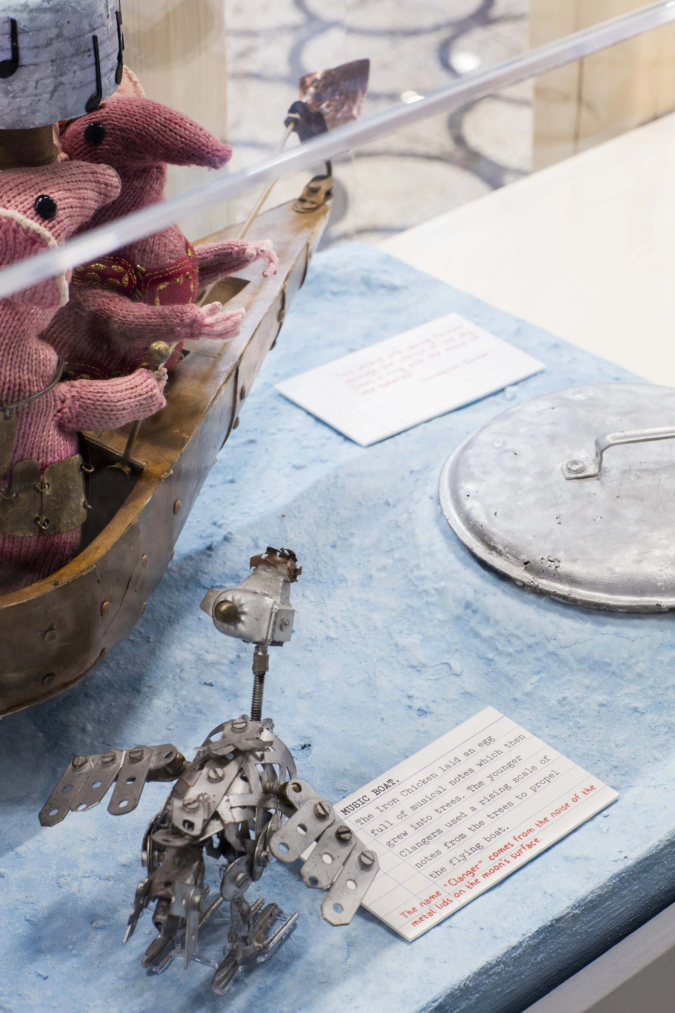 Two pink knitted mice on a wooden boat and a metal bird are standing on a rough blue base. An exhibit label next to them says “Music Boat” and is written with a typewriter, with red handwritten text beneath it.