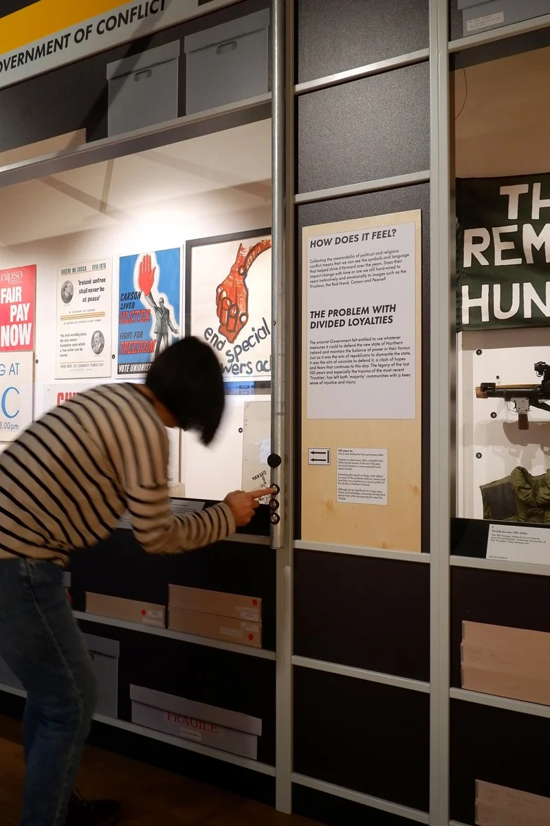 A visitor bends down to press buttons on a metal tube which sits to the side of a display of objects.