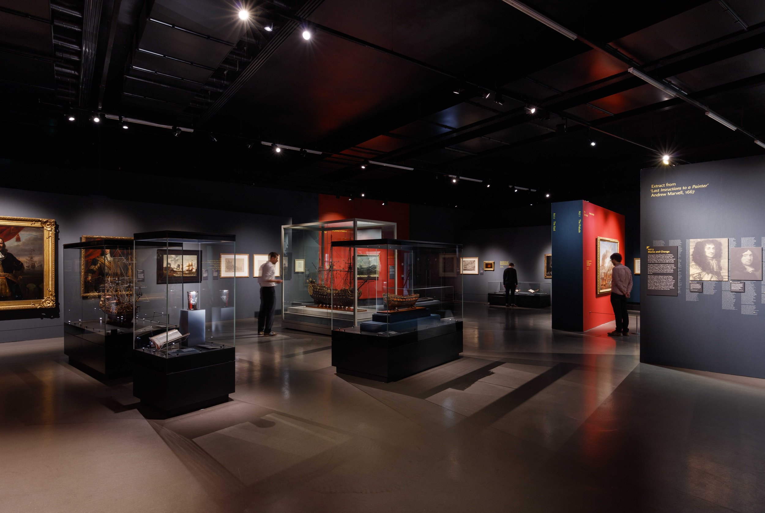 A low-lit gallery space with dark navy walls and red accent walls. Showcases in the centre of the room display large replica models of ships. Framed images on the walls include paintings of notable personalities and maritime scenes.