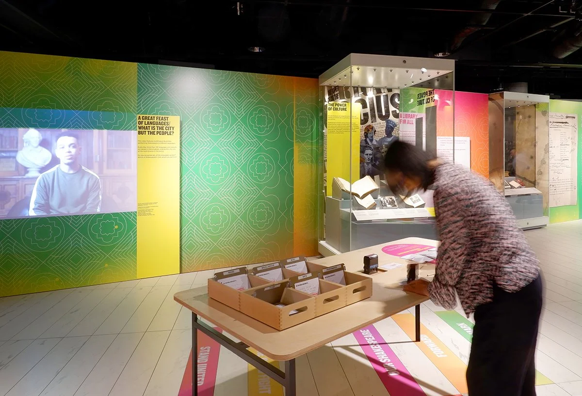 A woman stamps a card at a table in the middle of a colourful gallery. In the background a video projection of a man plays next to exhibition text asking "What is the city but the people?" To the right, showcases display open books.
