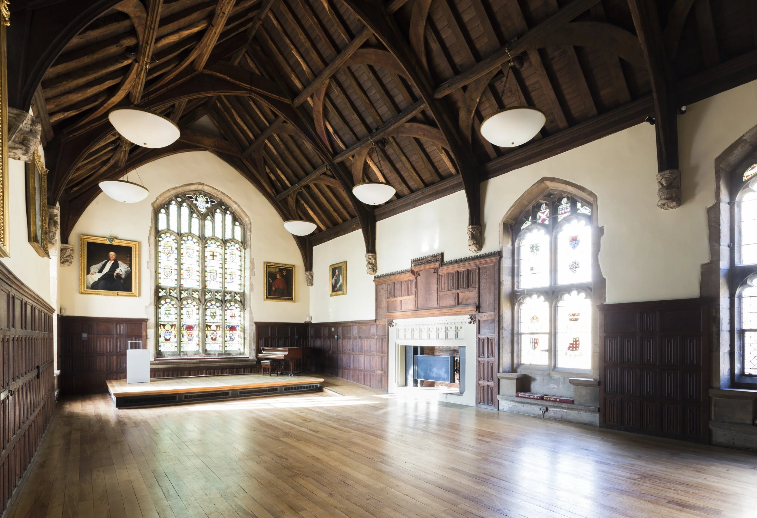 A hall with wood floor and vaulted ceiling, with a tall window at one end and two others on the adjacent wall, all with stained glass panels. The lower walls are wood panelled, with framed portraits above them.