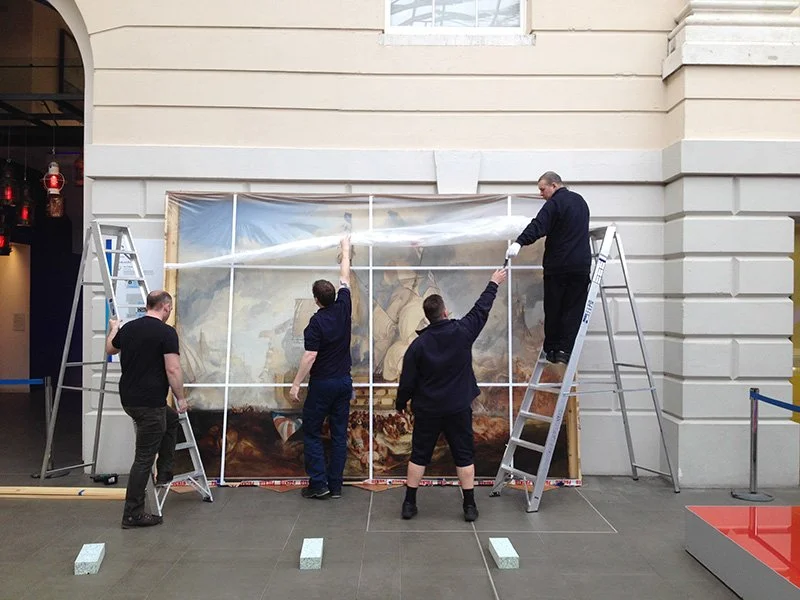 Four men dressed in black stand by a Turner's Battle of Trafalgar painting, which is packaged and leaning against an outdoor wall. Two of the men are standing on ladders removing the packaging.