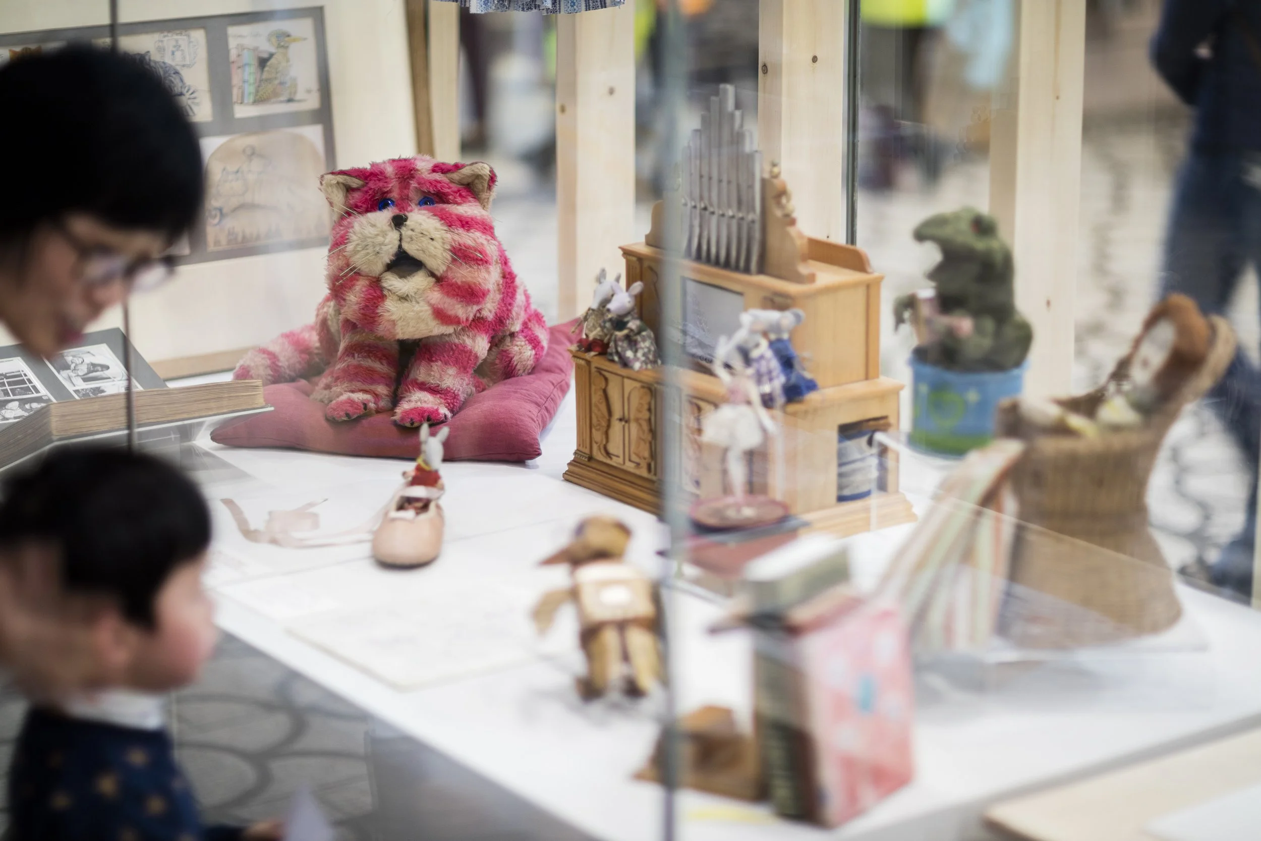 An adult and child look at a showcase with various toys inside, including a large pink fluffy cat, a mouse in a ballet shoem mice standing on a model of a church organ, a wooden bird, and a doll in a rocking chair.