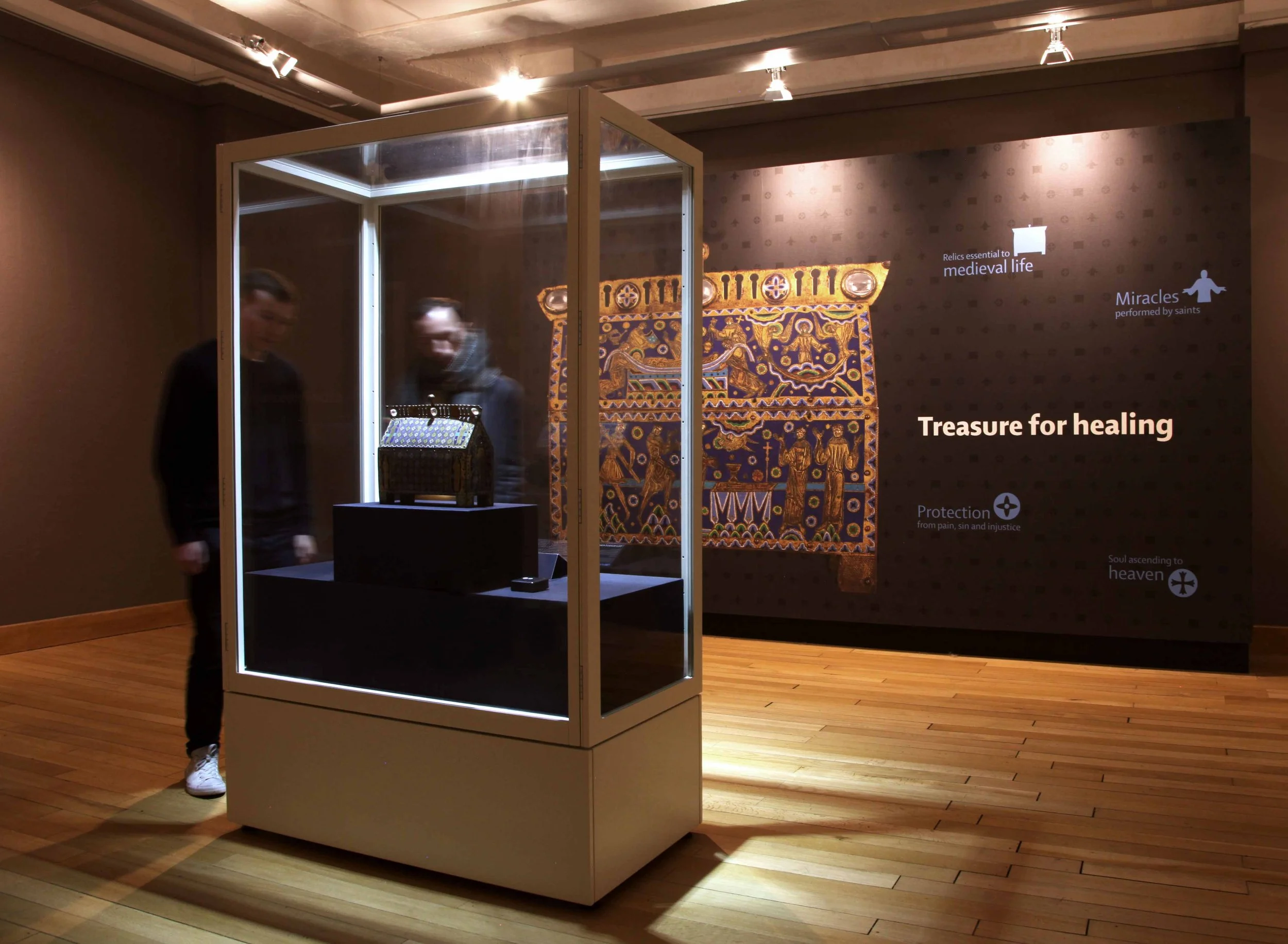 Two men look at a spotlit object in a showcase in a gallery. Behind them is a wall-sized image of the object – an ornate painted box with human figures – and the title Treasure for Healing.