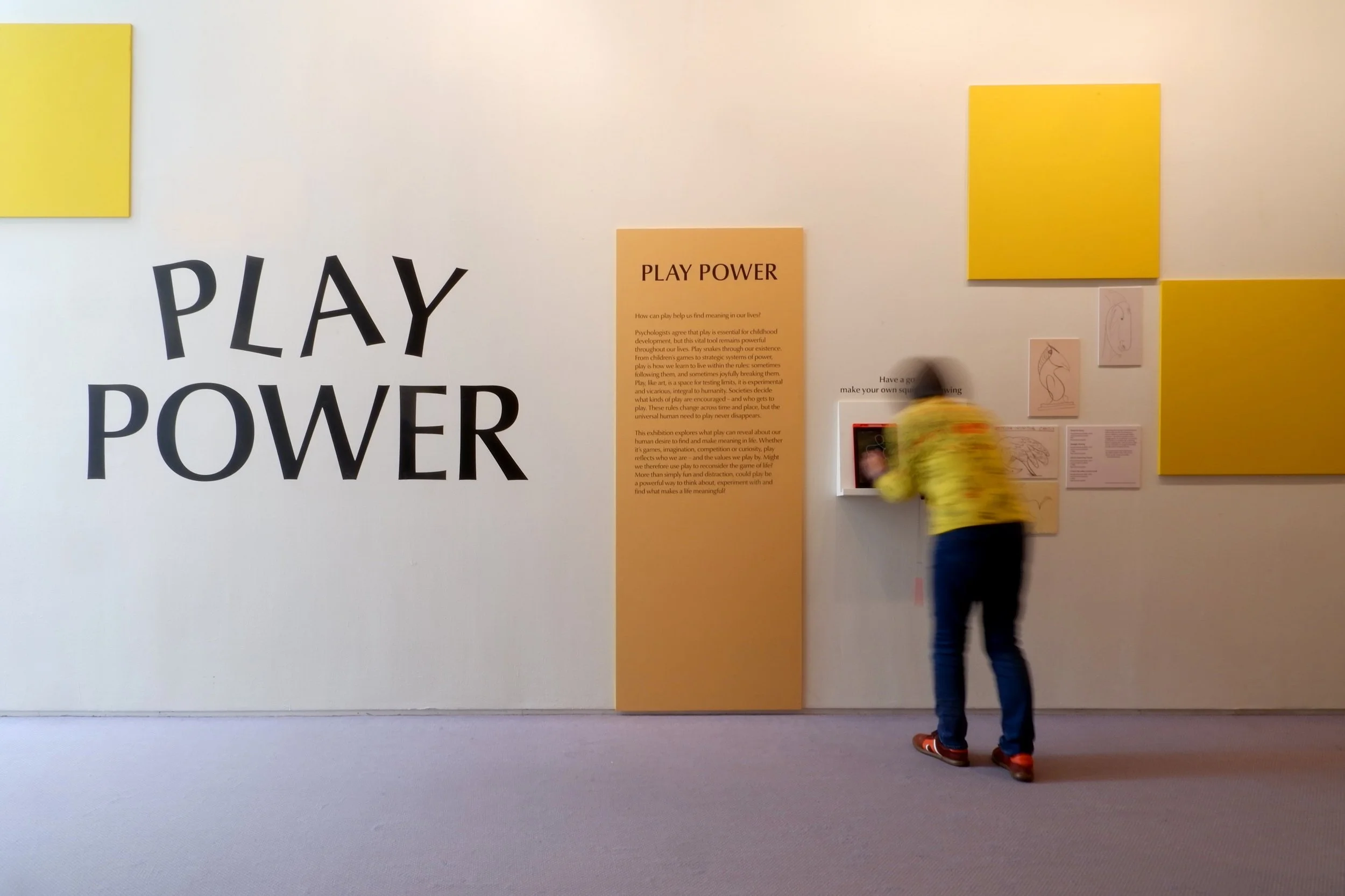 Gallery wall with graphic panel and exhibition title reading "Play Power" as graphic stencilled on the wall. Woman doing a doodling interactivity.