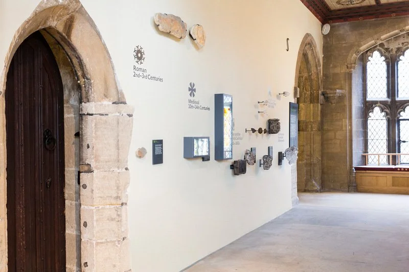 The white interior wall of a church leading to a large window. To the left is a wooden door. The arch around it shows original stonework with text reading “Roman 2nd-3rd Centuries.” Stone artefacts, text panels and lightboxes are mounted to the wall.