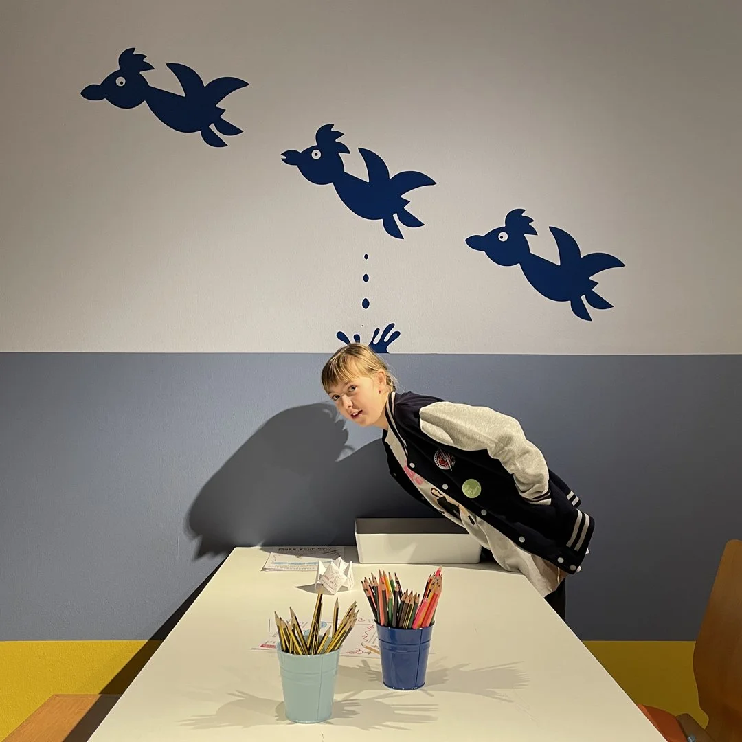 Two people look at books displayed in green showcases in front of a wall decorated with repeating green motifs. Along the wall are framed illustrations and large cardboard cut-outs of palm trees which hang over the exhibits.
