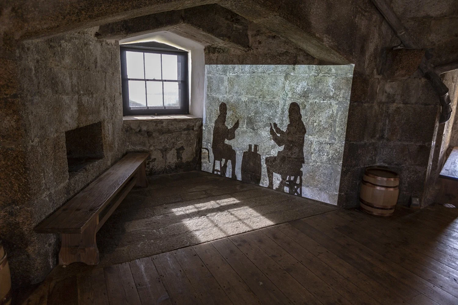 A room with rough stone walls and a small window letting in natural light. On one wall is a projection of the silhouettes of two seated male figures sharing a drink around a barrel. Opposite the projection is a long wooden bench for visitors to sit.