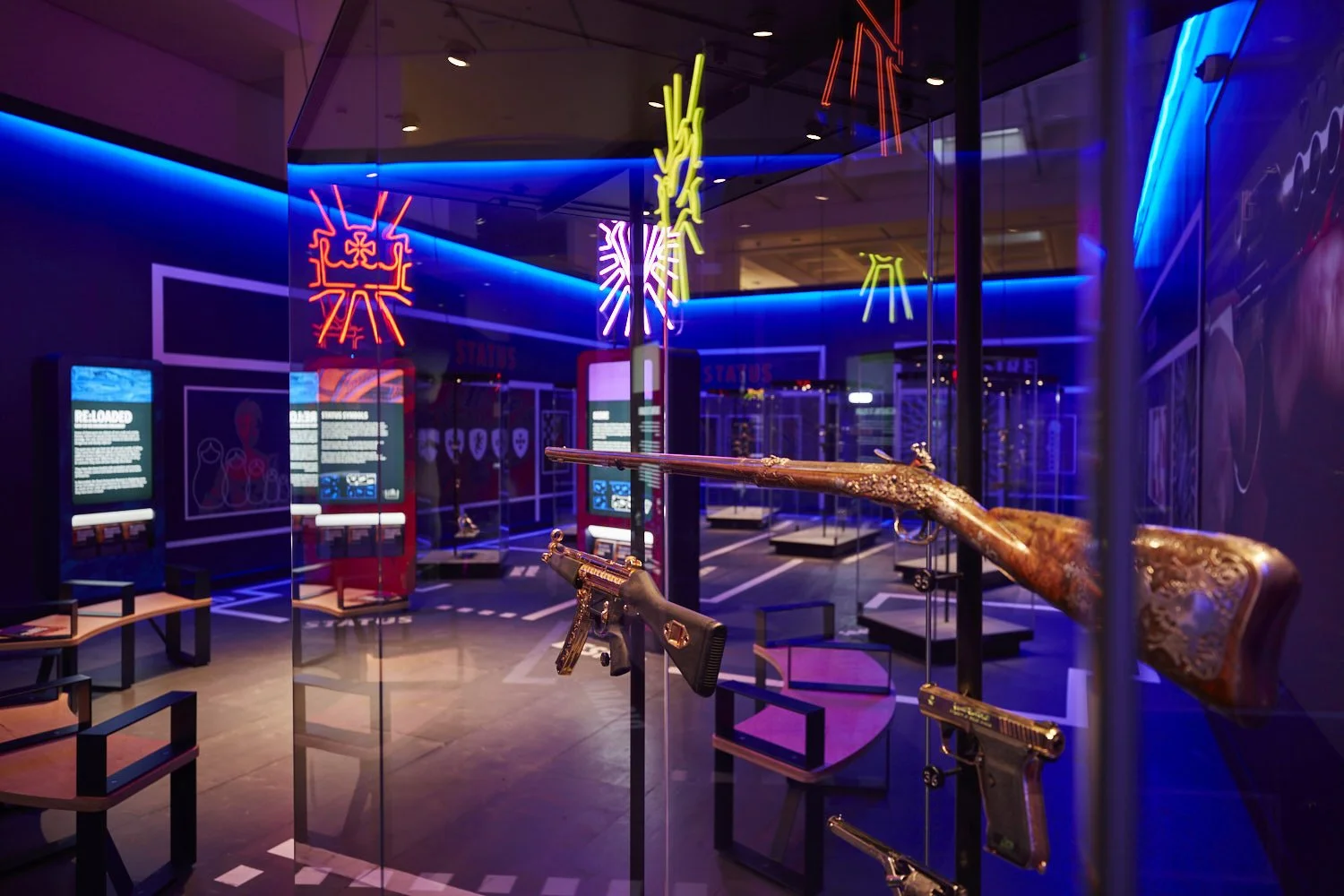 A view of the temporary gallery. In the background are three walls with top-level blue lights illuminating graphic surfaces. In the foreground is a showcase with guns mounted on thin rods so they appear to float in space.