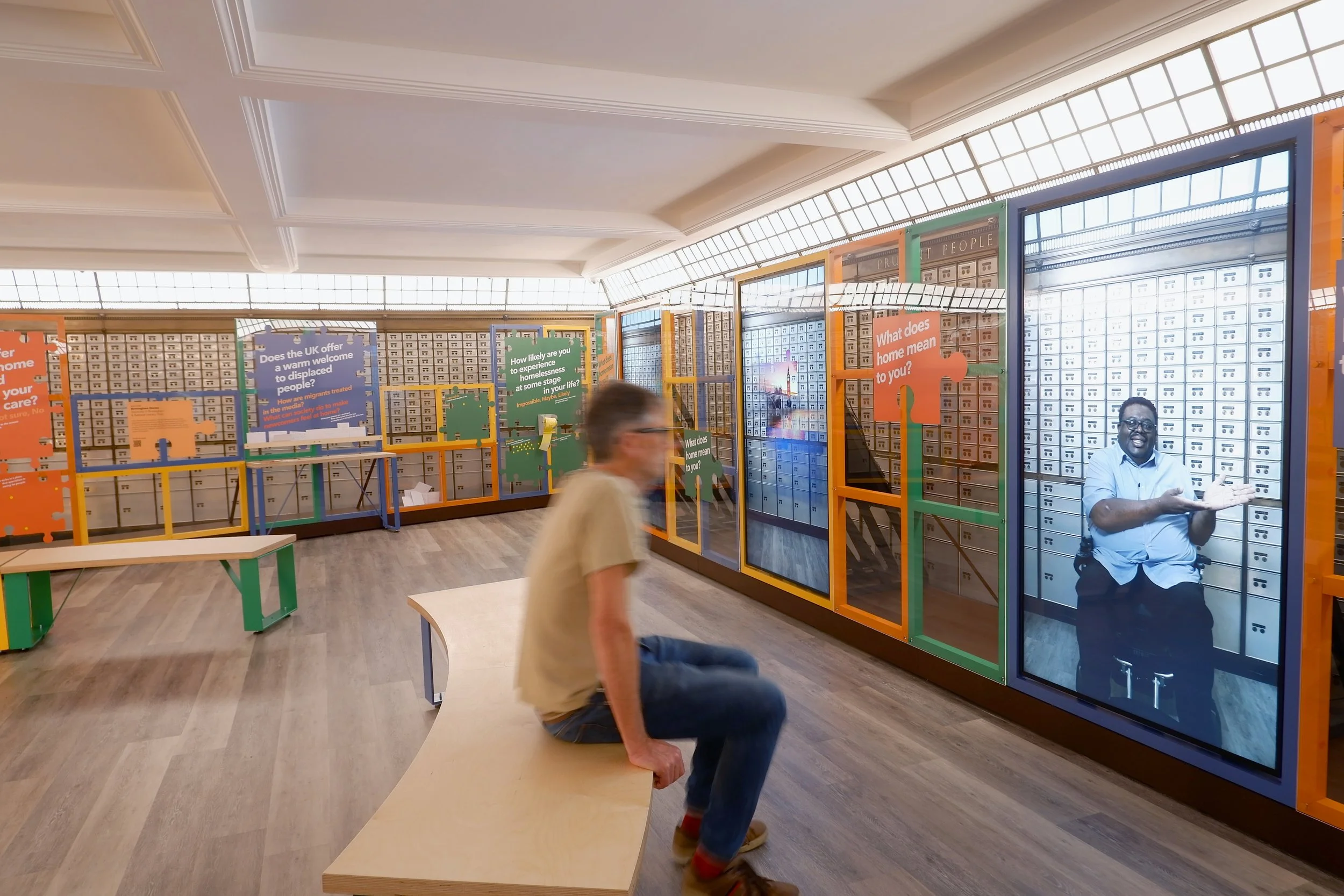 A man sits on a wooden bench opposite a large video screen of a smiling man in a wheelchair. It is one of three screens connected by a colourful structure that extends around the room. In the background, display panels ask questions about home.