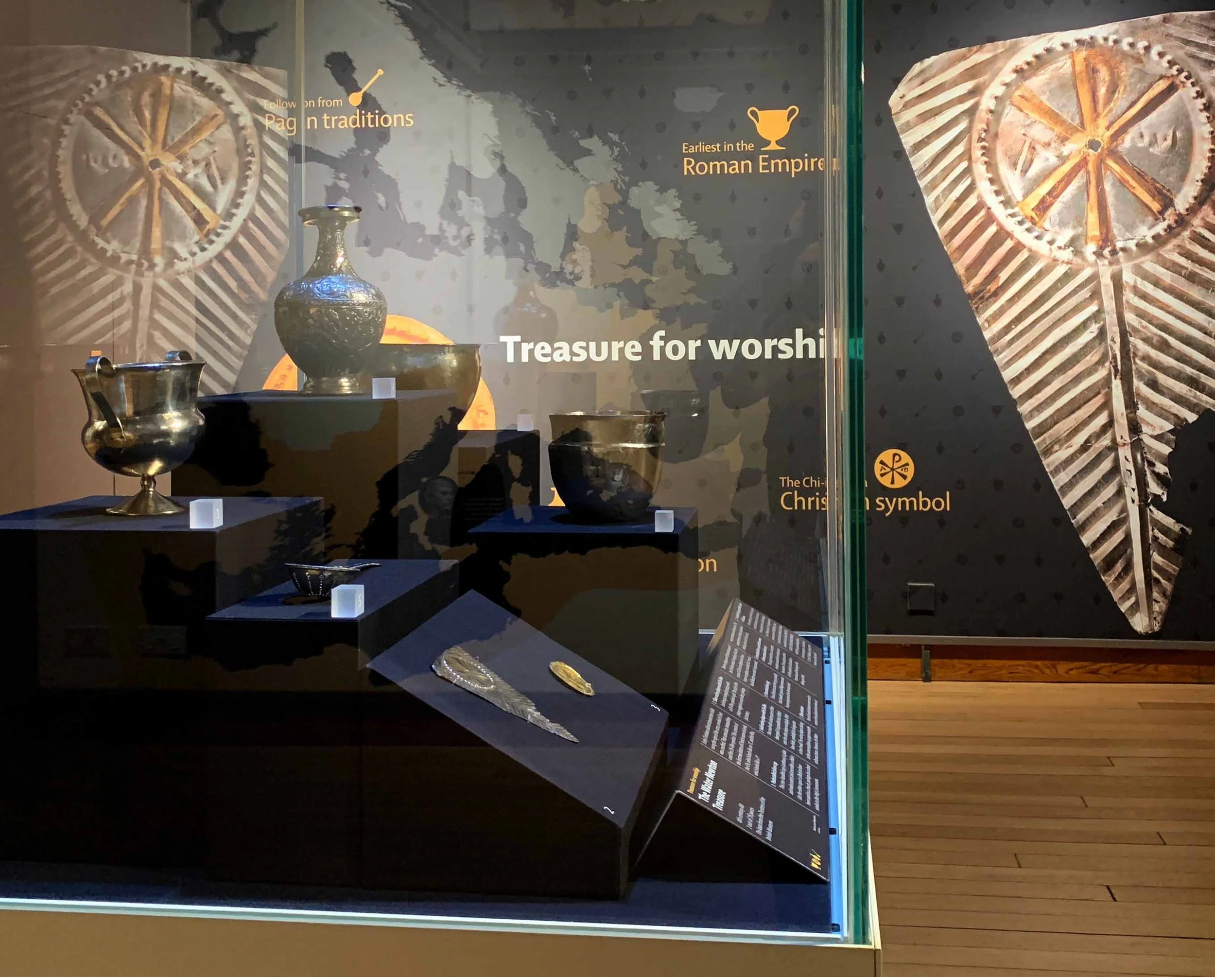 A selection of metal objects in a showcase, including a vase and goblet. Behind it, a wall graphic shows a close-up image of a triangular metal plate with a circular Christian symbol. The title reads "Treasure for worship".