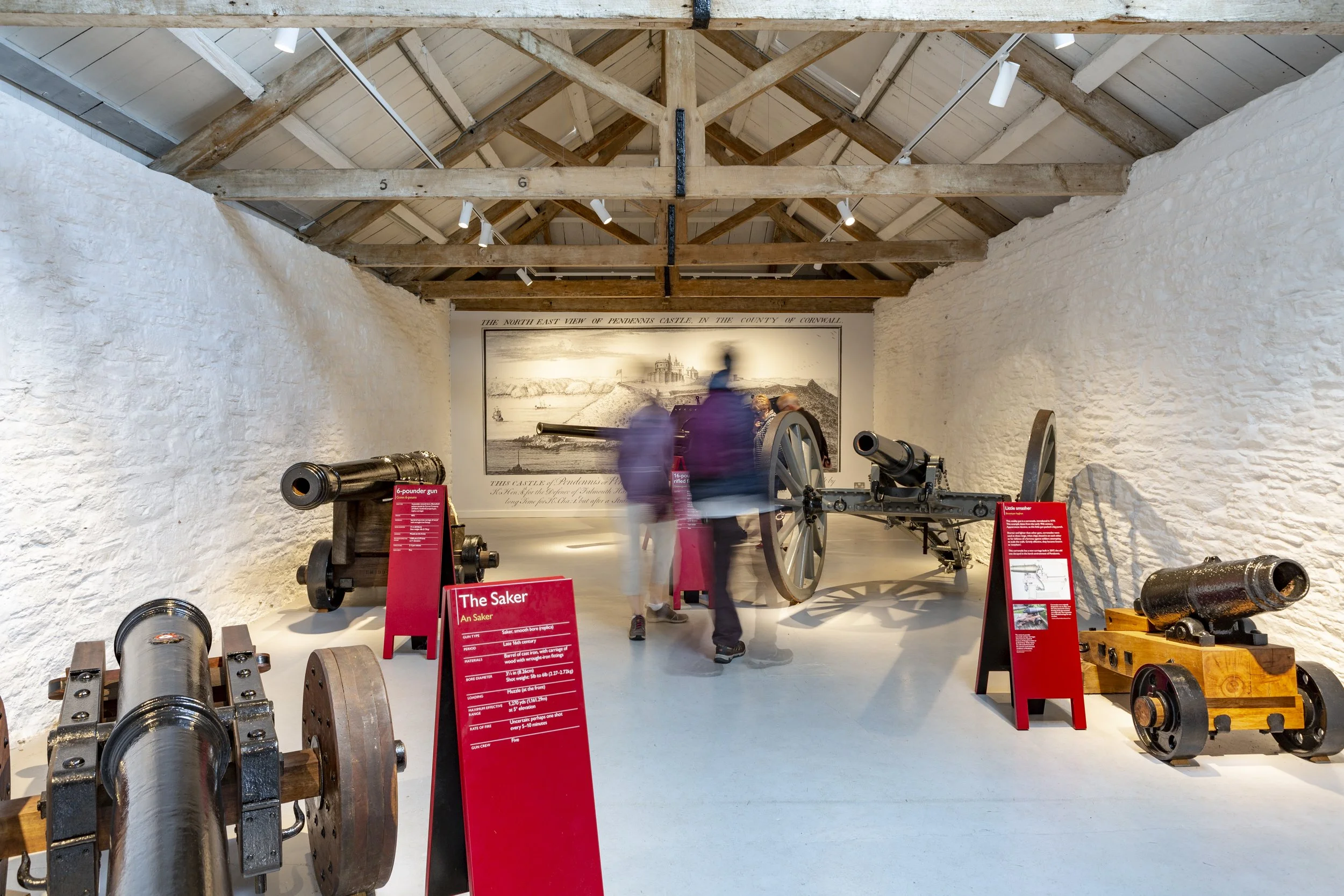 Visitors in a white-walled stone room with several cannons on display and a large illustration of Pendennis Castle on the back wall. Red A-frame text panels provide further information about each gun.
