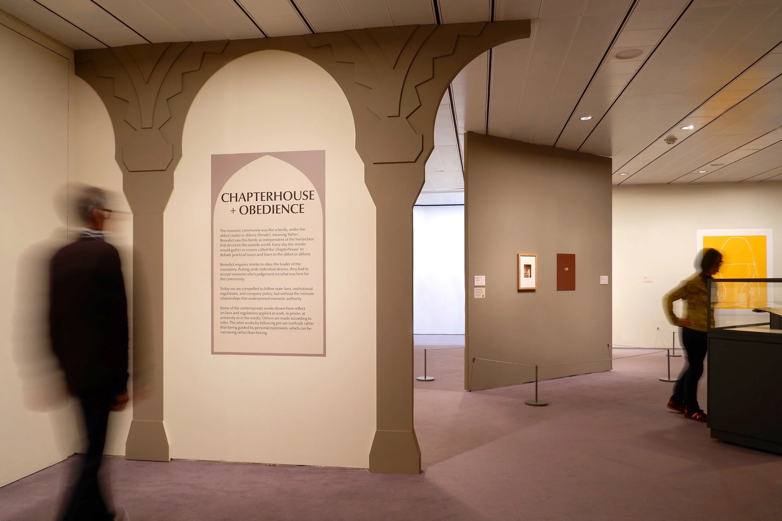 Man standing in front of text panel in gallery space. Decorative architectural elements on the wall representing abtract design of medieval cloisters. Floating division wall in the background with works of contemporary art.