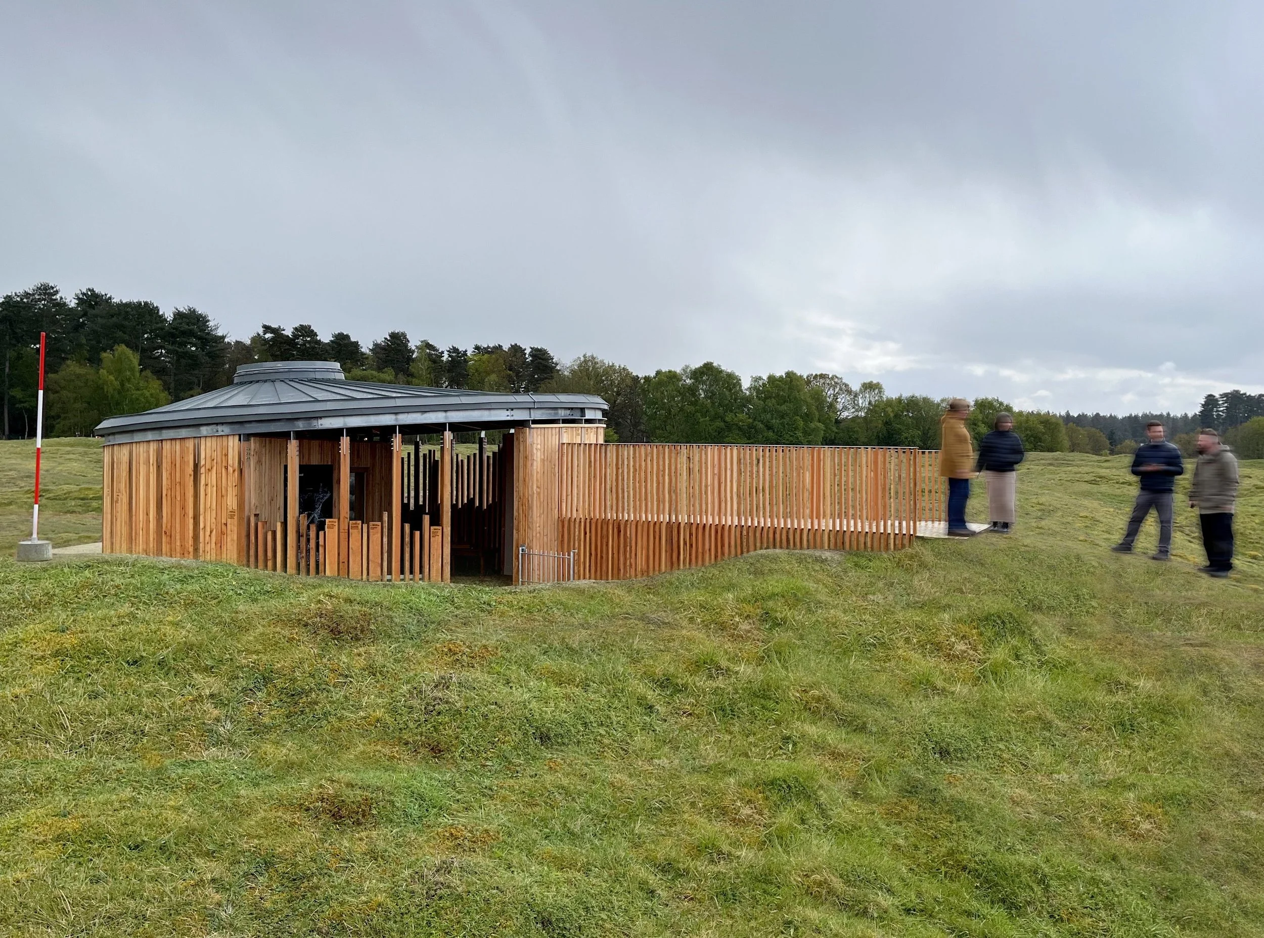 At Grime's Graves in Norfolk, a wooden structure in a field acts as an information pavilion with a ramp, drawing a crowd of people. It functions as an info pavilion with a ramp. The round structure attracts a group of people.
