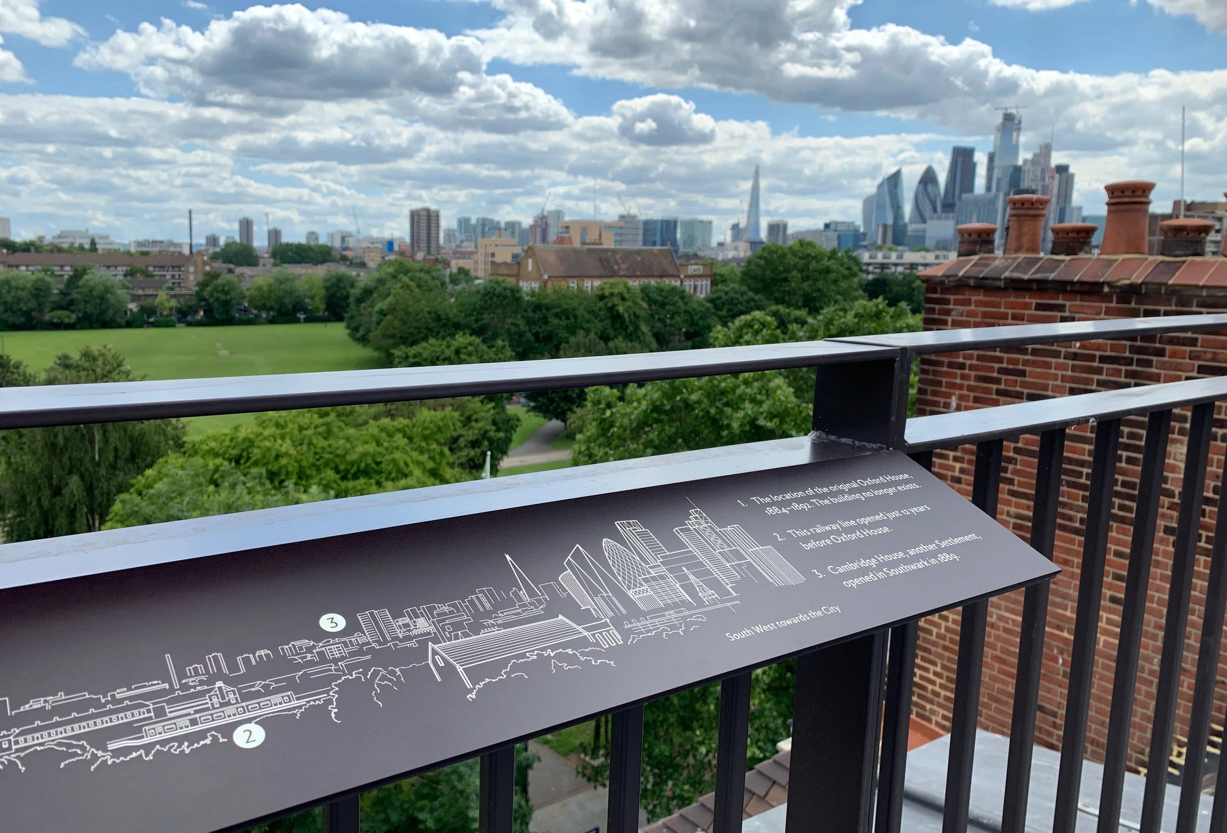 View of the London skyline from a roof, with a green park and skyscrapers in the distance. Affixed to the railing is a map of the skyline with white line illustrations of key buildings and locations.