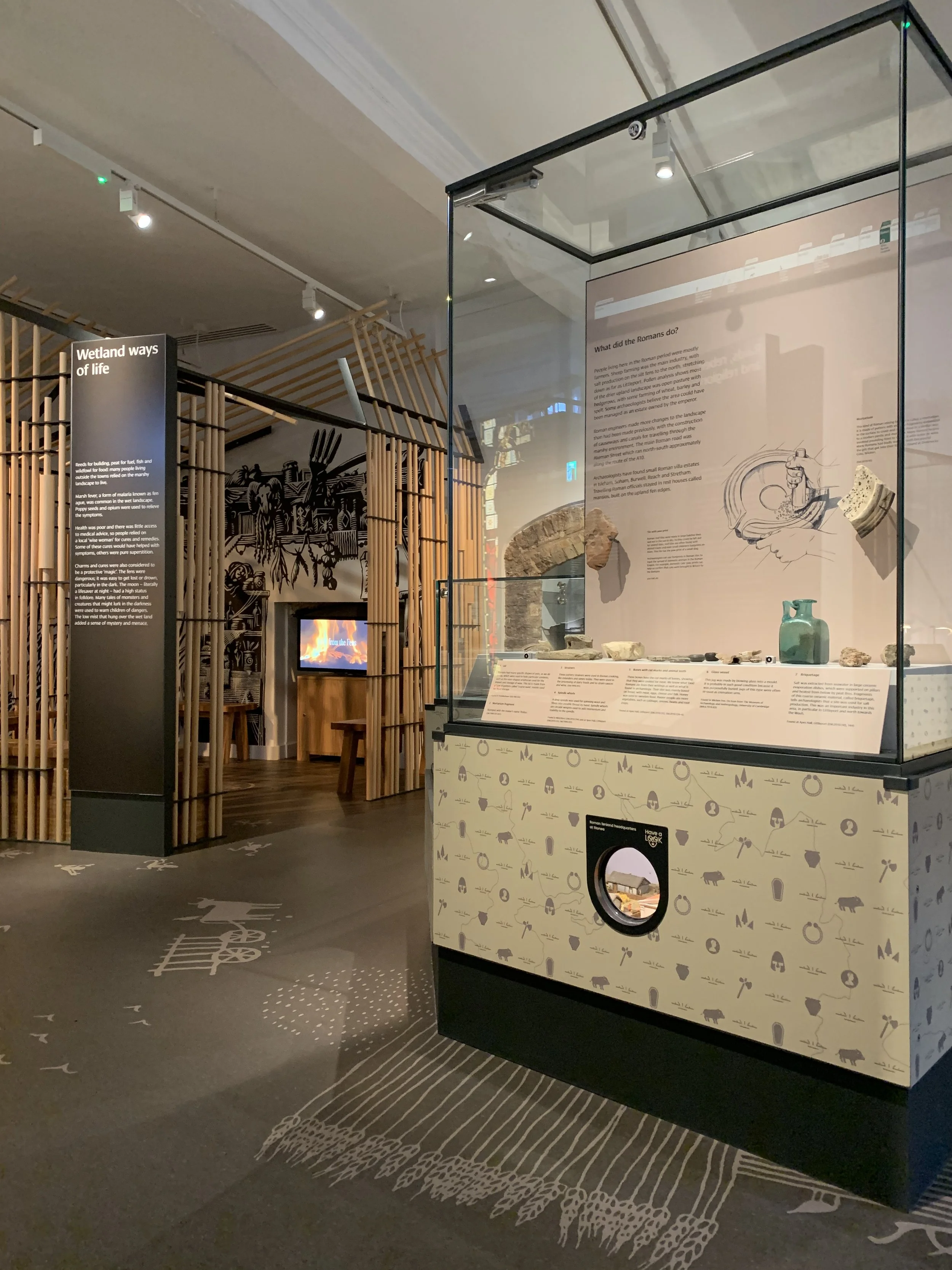 A sand-coloured showcase in a gallery. It has a peephole in its base and archaeological artefacts on display. The gallery floor is decorated with agricultural motifs.