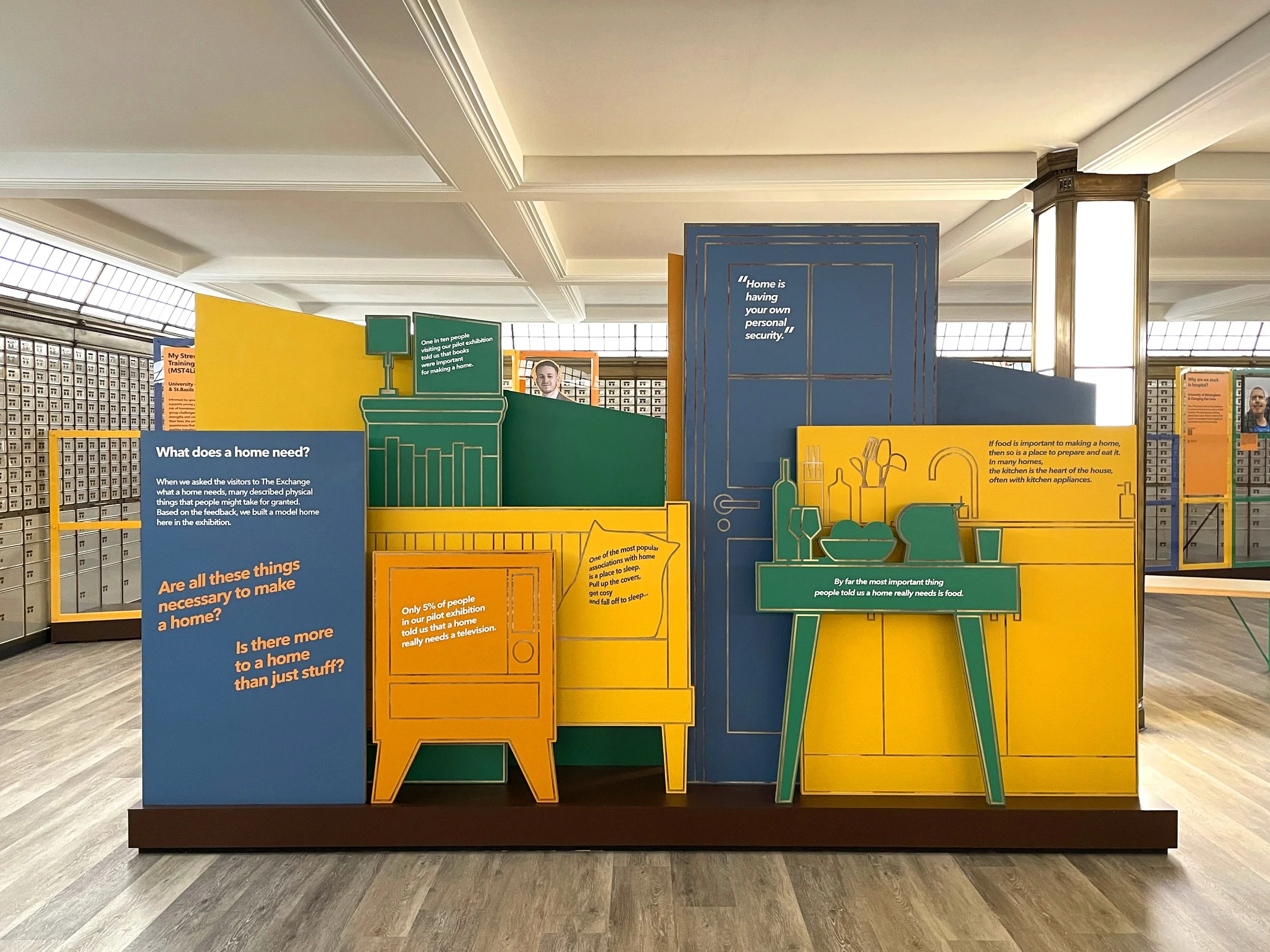 A large display of blue, orange, yellow and green 2-dimensional shapes in the centre of a room. Some are cut in the shapes of domestic furniture, like a TV set, sofa, door and kitchen sink, and feature quotes and questions about what home is.