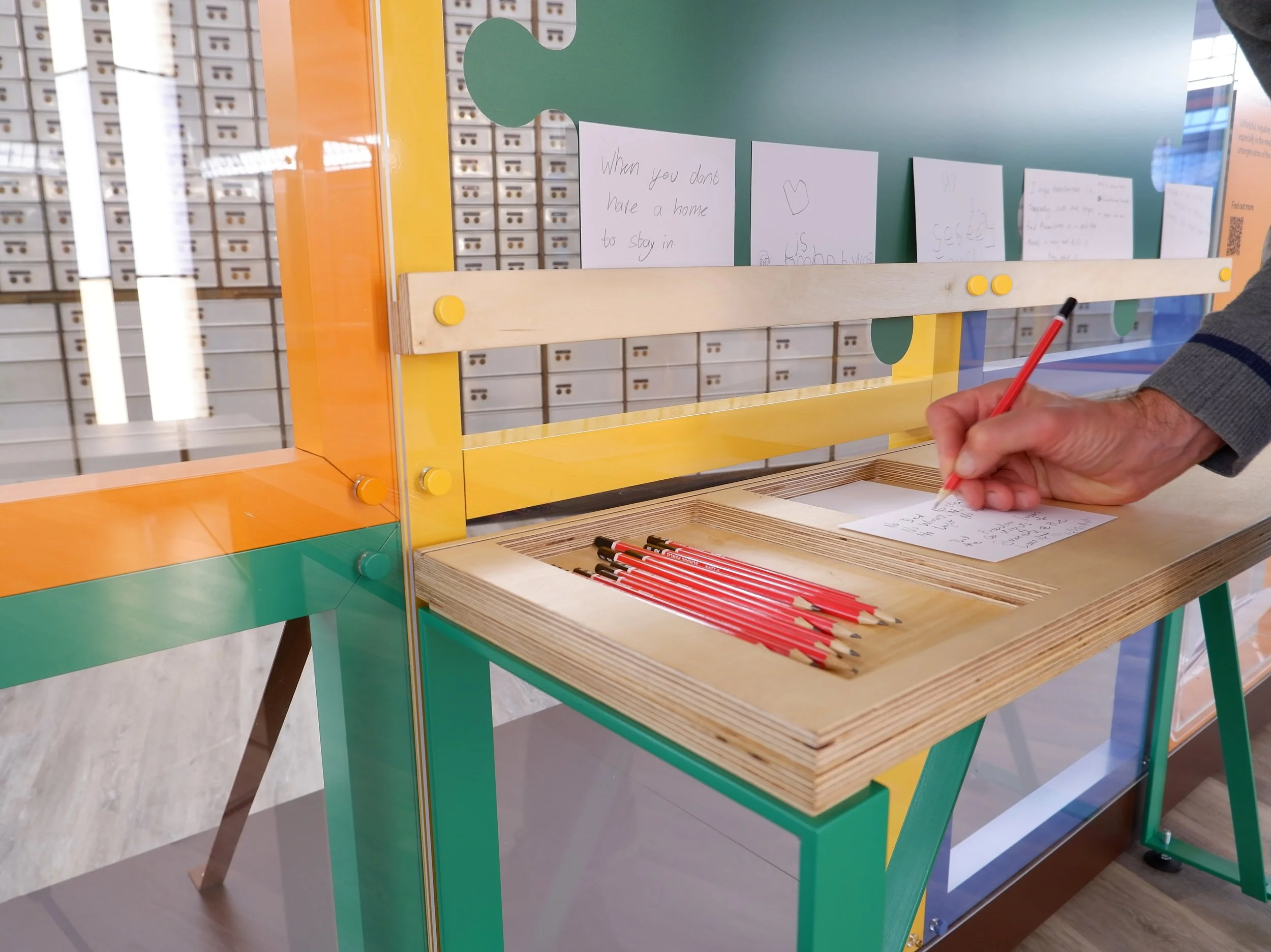 A person writes on a postcard with a red pencil on a wooden ledge attached to a colourful yellow, orange and green exhibition structure. Above the ledge, other handwritten postcards are displayed. One reads “when you don’t have a home to stay in”