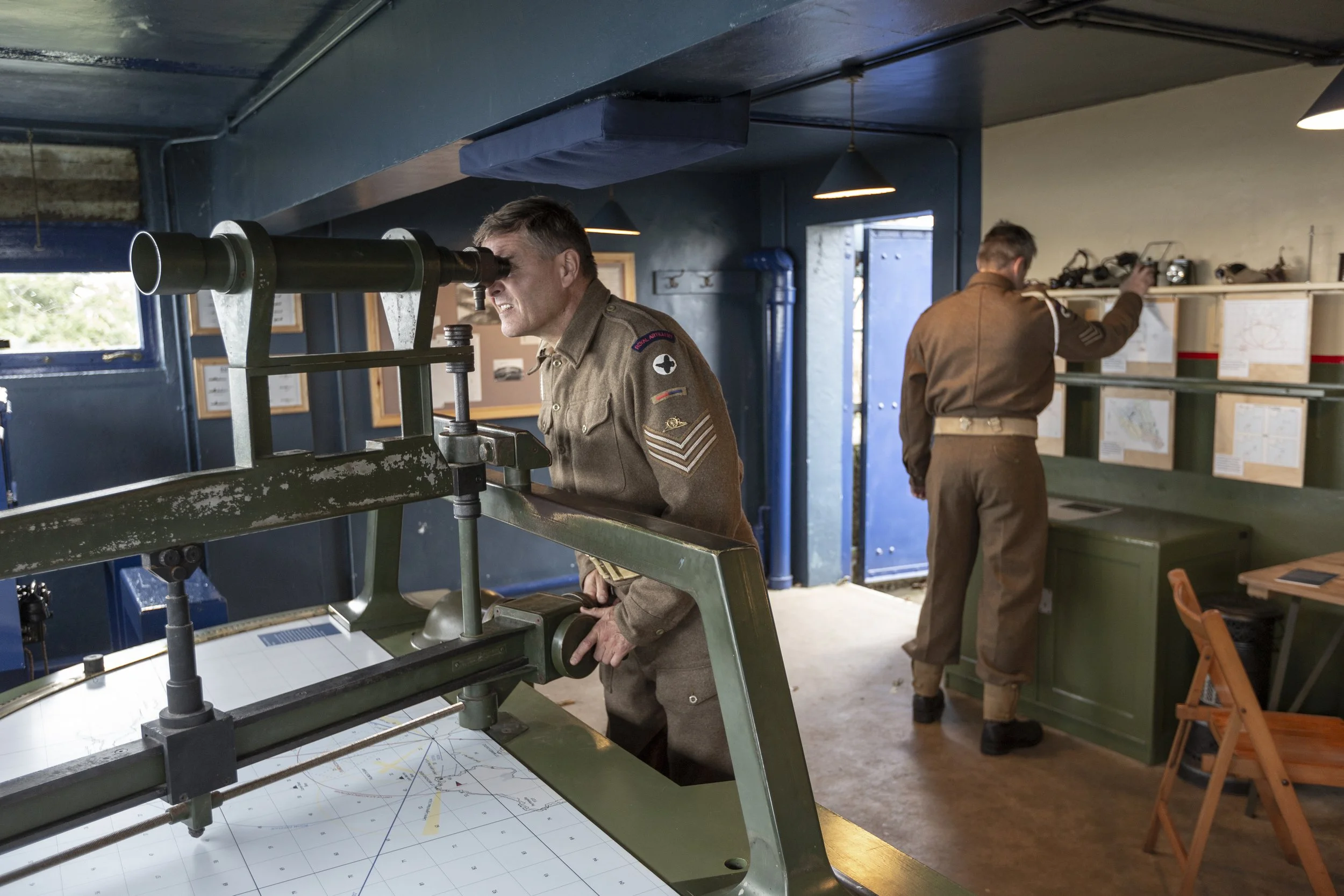 Two men in military uniforms use technical equipment in a room. One looks through a scope with a plotter and map beneath it.