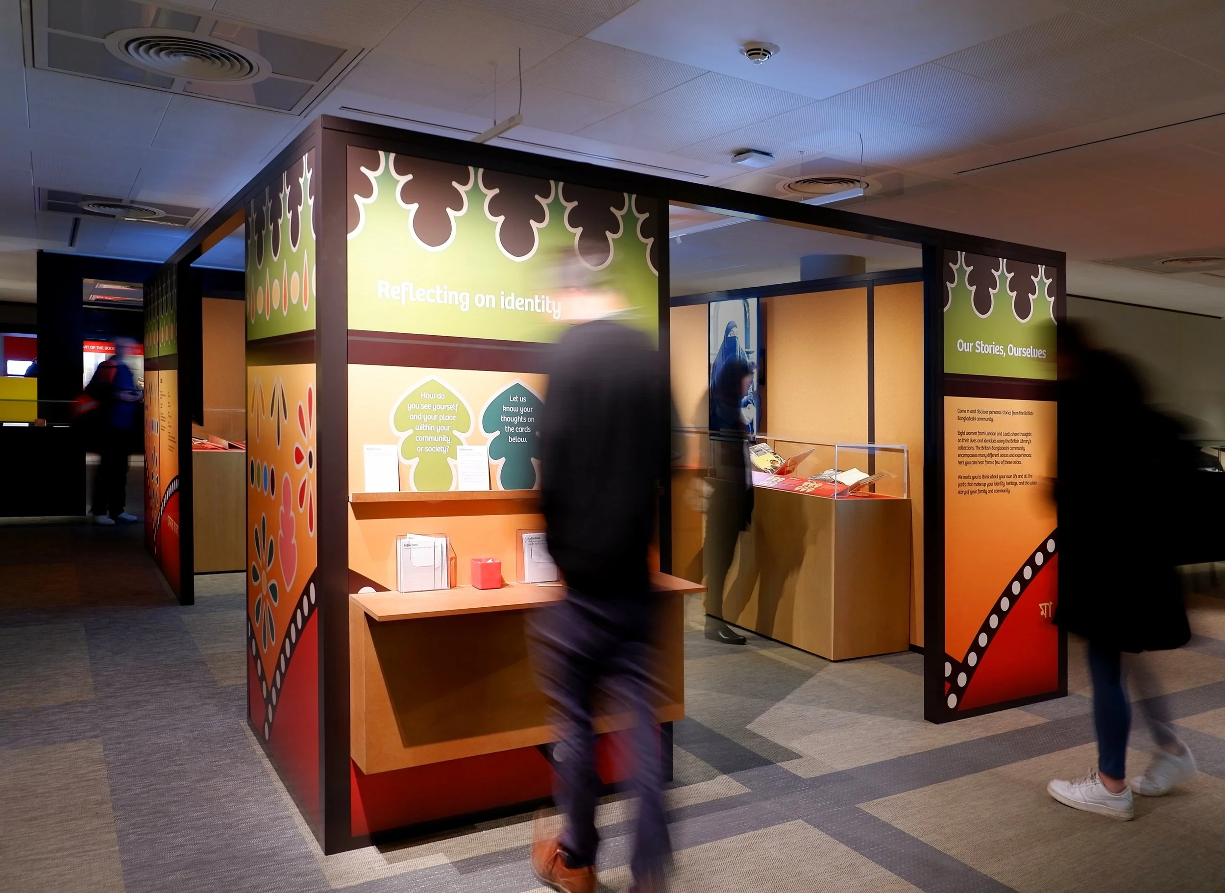 A colourful rectangular structure standing in the middle of a gallery at the British Library. The graphic surfaces of the structure are colourful and include shapes of flowers and other symbols related to Bangladeshi culture.