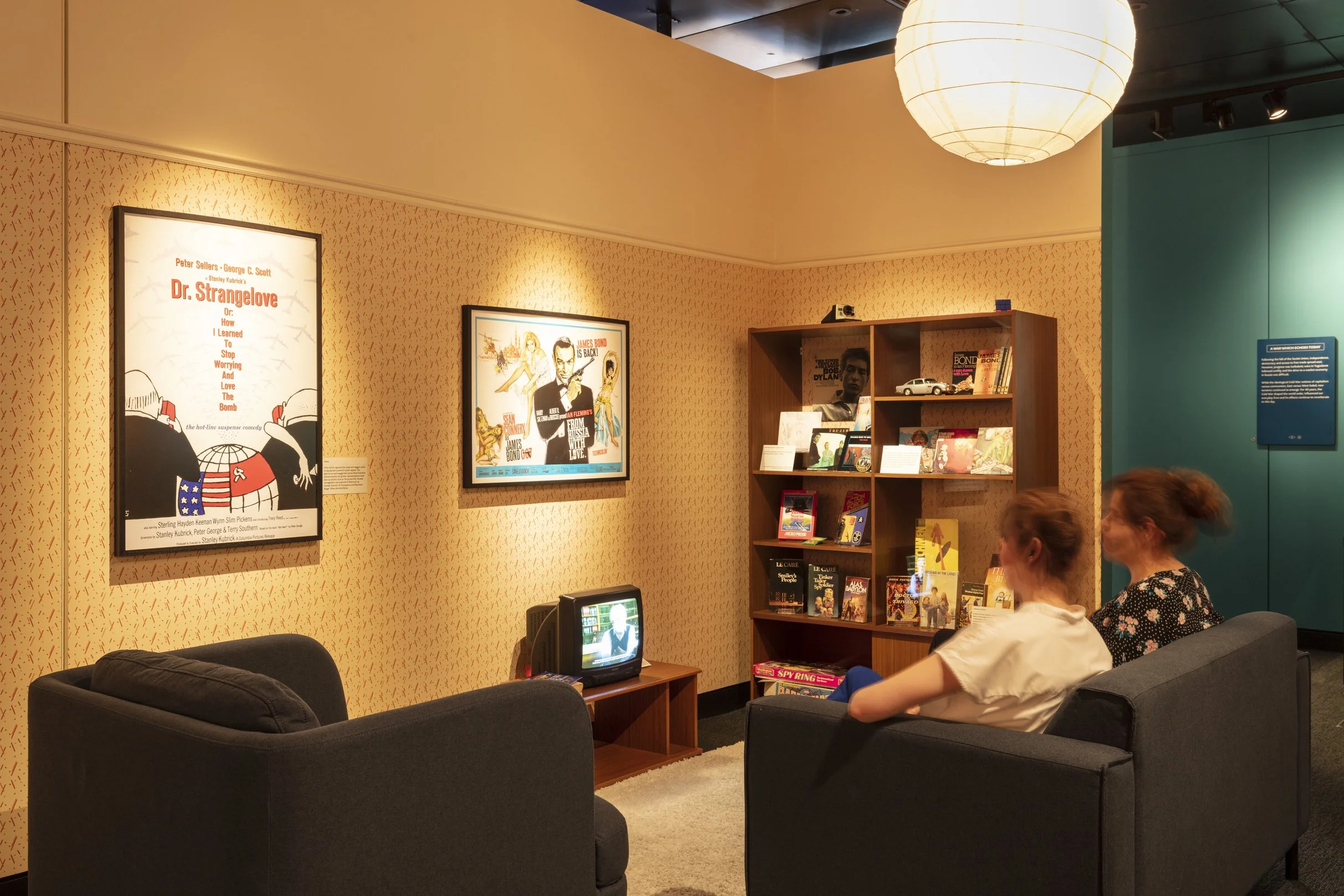 Two women on a sofa face a TV in an exhibition that looks like a living room with yellow wallpaper. Above the TV are posters for the films Dr Strangelove and From Russia With Love. A bookshelf displays related pop culture paraphernalia.