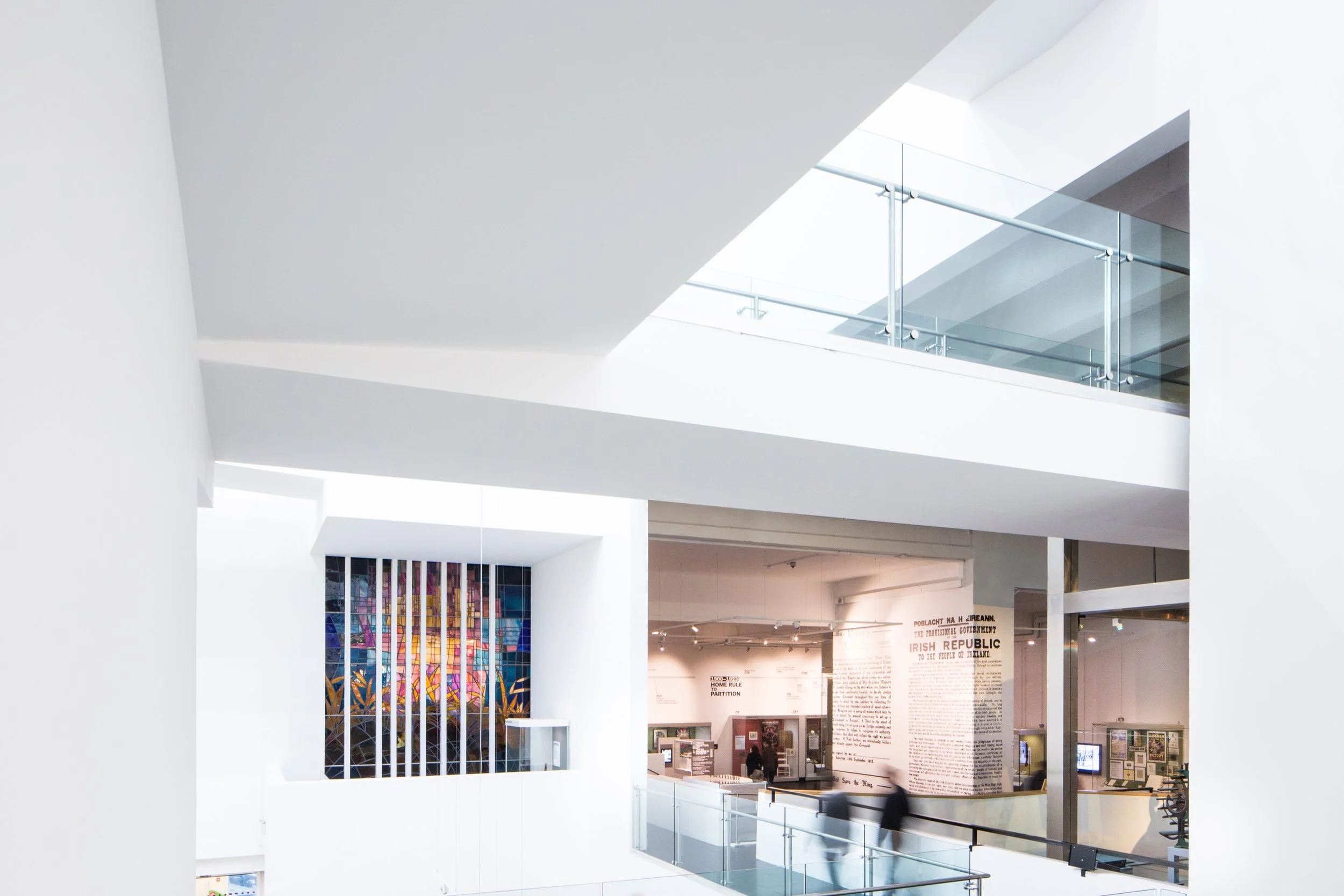 A white building interior with multiple levels. Visitors walk towards a gallery with showcases and exhibits in the background. On the left a blue and orange stained glass window is set in a white wall. Above them is a walkway on the second floor.