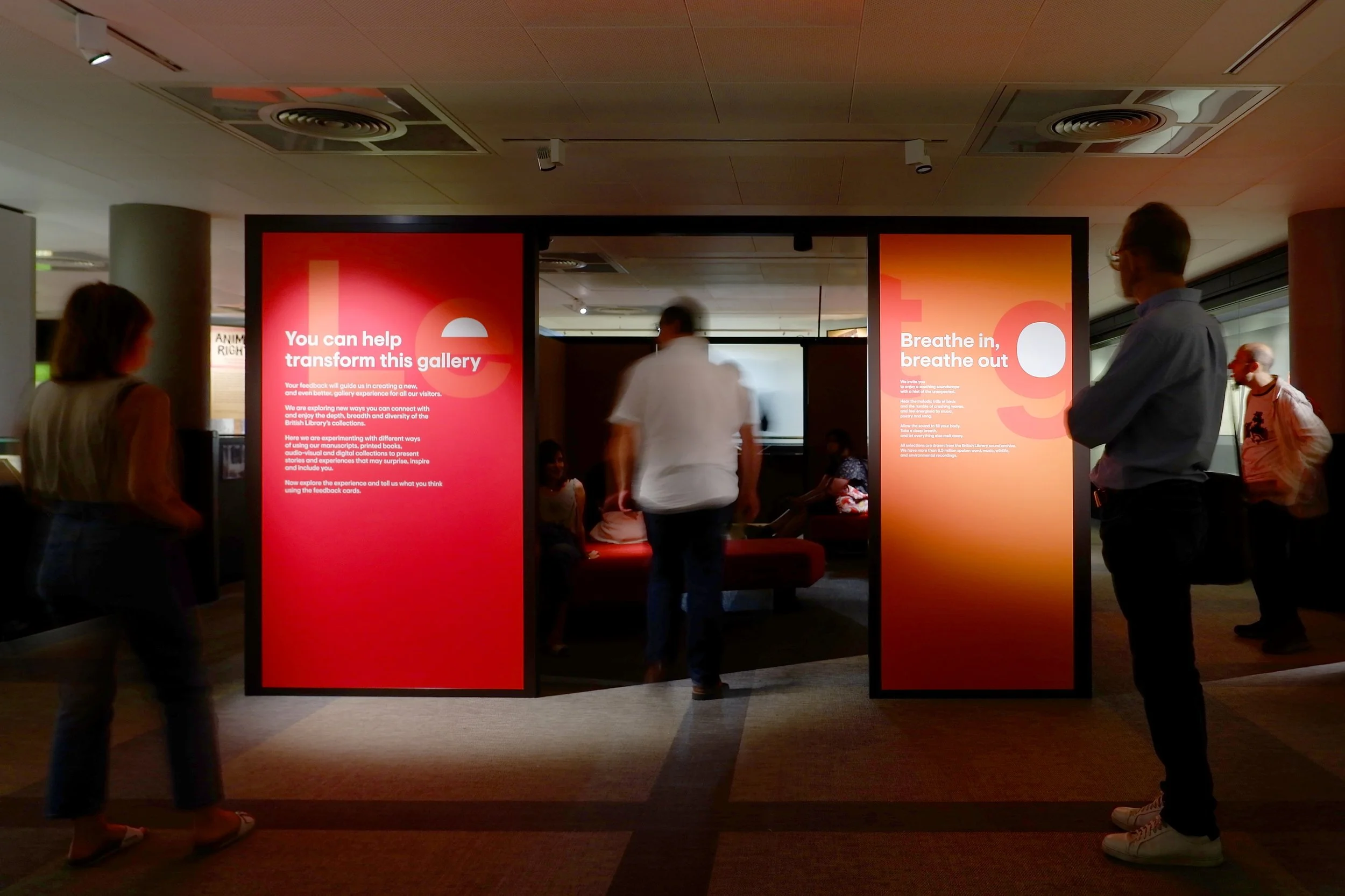 A rectangular structure stands in the middle of a gallery at the British Library. The graphic panels are red and carry textual information. A man enters the structure while several people sit on the benches inside.
