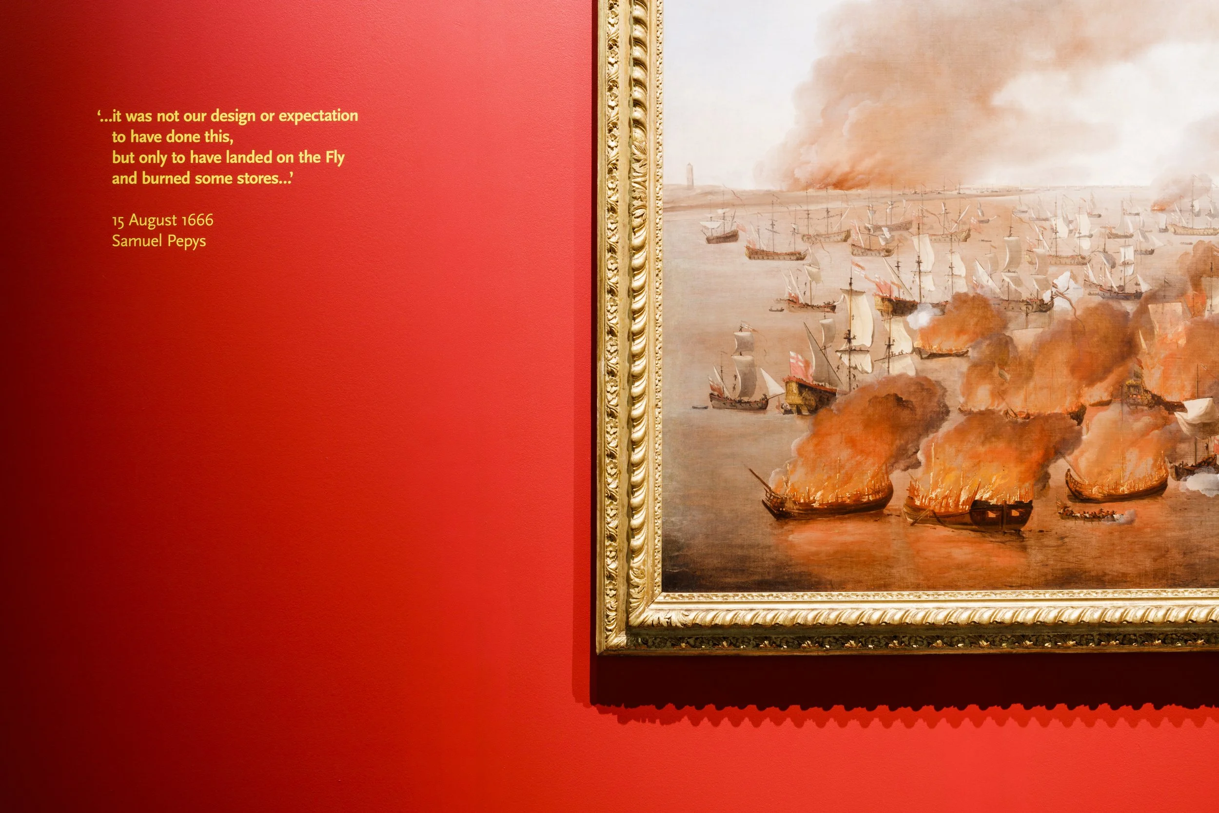 A red wall and the bottom left corner of a framed painting of a fleet of ships on fire. A 1666 Samuel Pepys quote on the wall reads: “it was not our design or expectation to have done this but only to have landed on the Fly and burned some stores.”