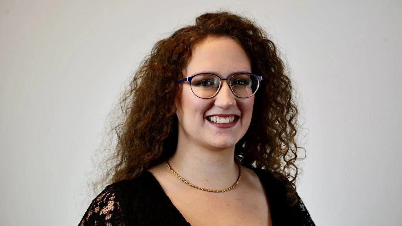 Woman with curly hair wearing glasses and a black top, smiling against a plain background.