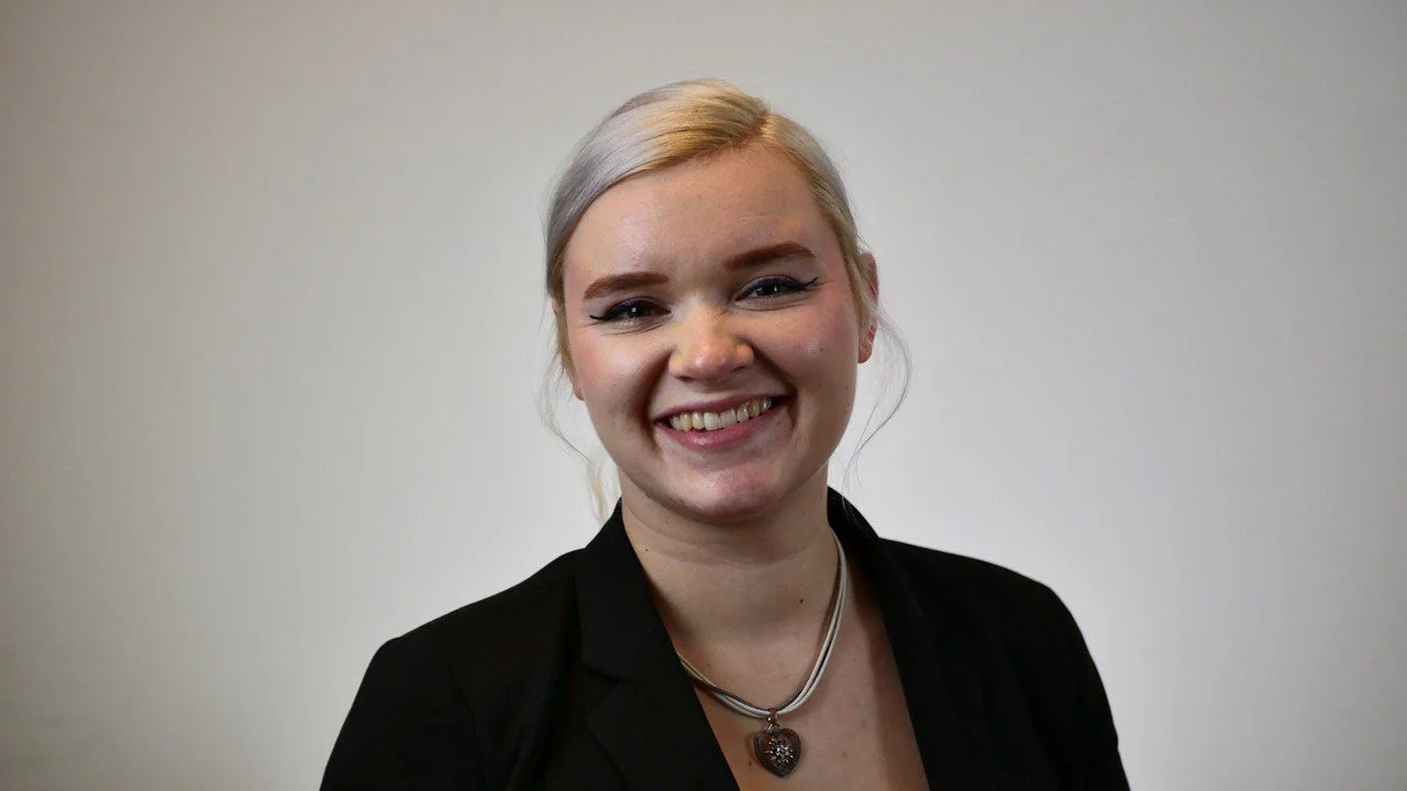Smiling person with blonde hair, wearing a black jacket and necklace, against a plain background.