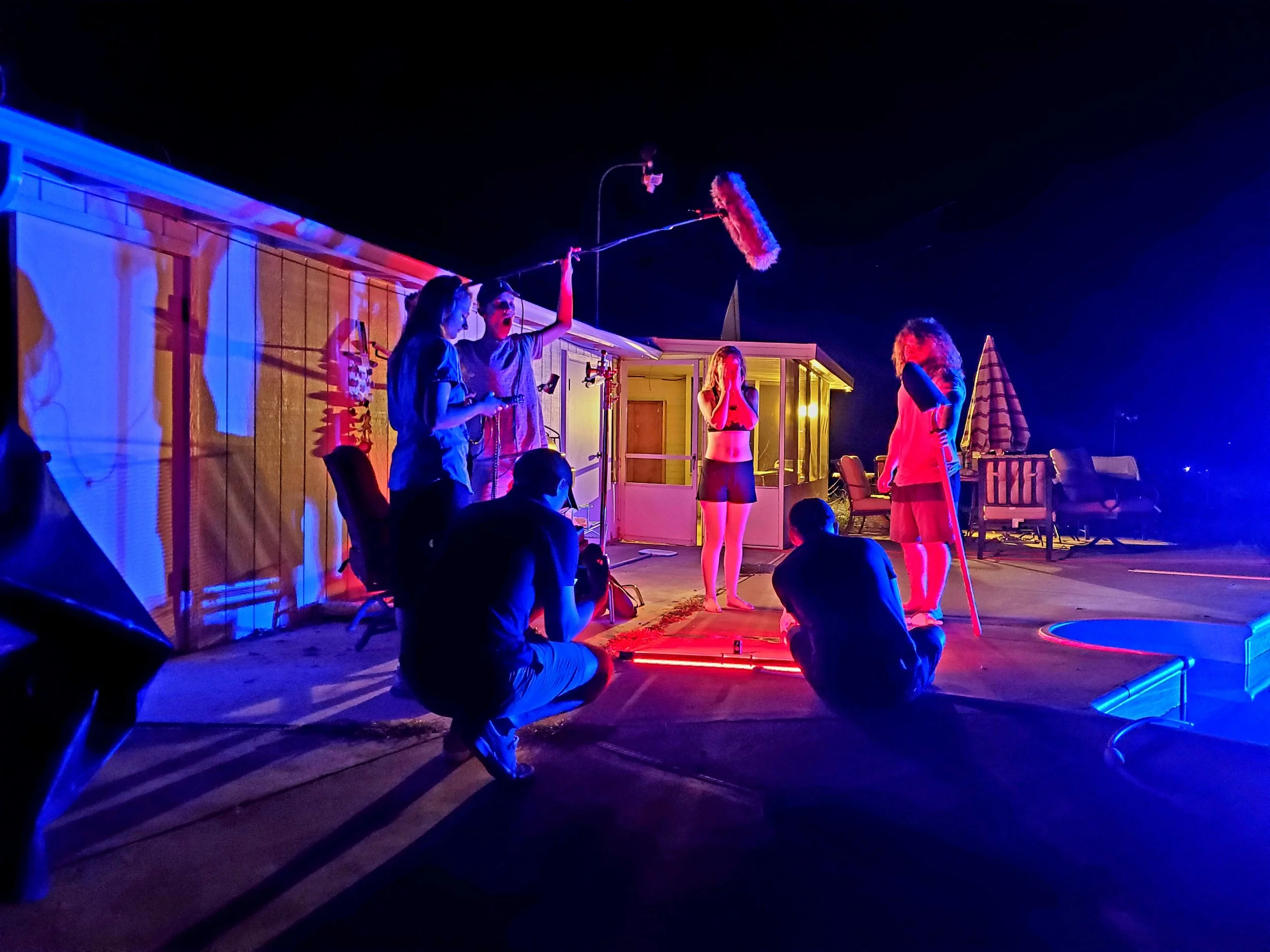 A nighttime film set scene with a crew filming outside a house. Several people are involved in the process, including a cameraman, a boom operator, and a director. One person is in costume, possibly acting, while others assist with equipment. Bright blue and red lights illuminate the area, creating dramatic shadows on the walls.