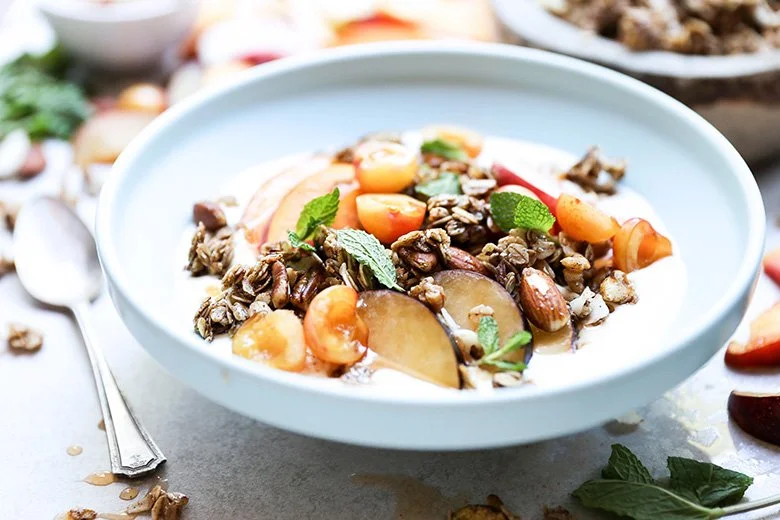 A bowl of yogurt topped with granola, fresh peaches, strawberries, and mint leaves.